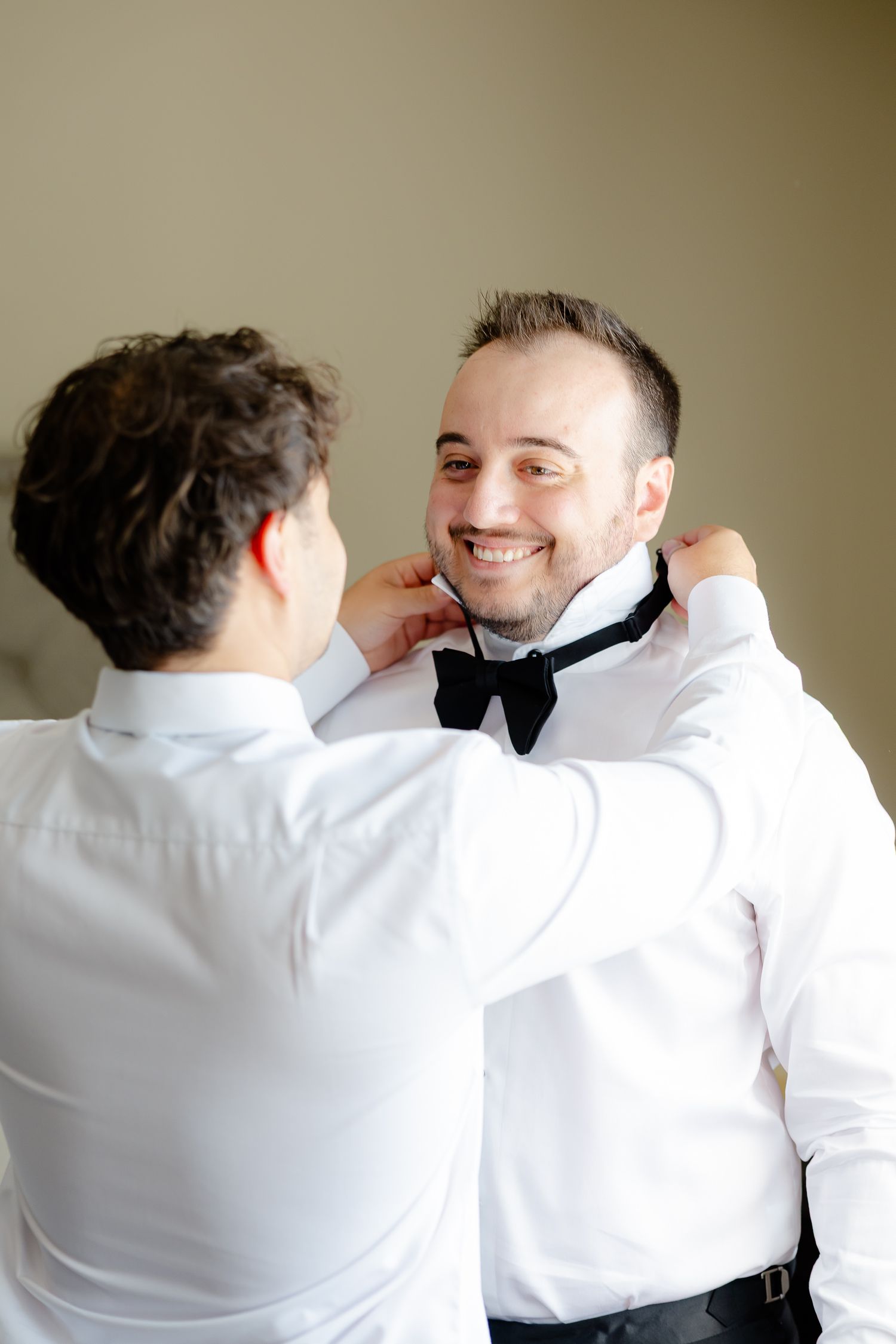 Two men in formal white shirts and bowties share a lighthearted moment while getting dressed.