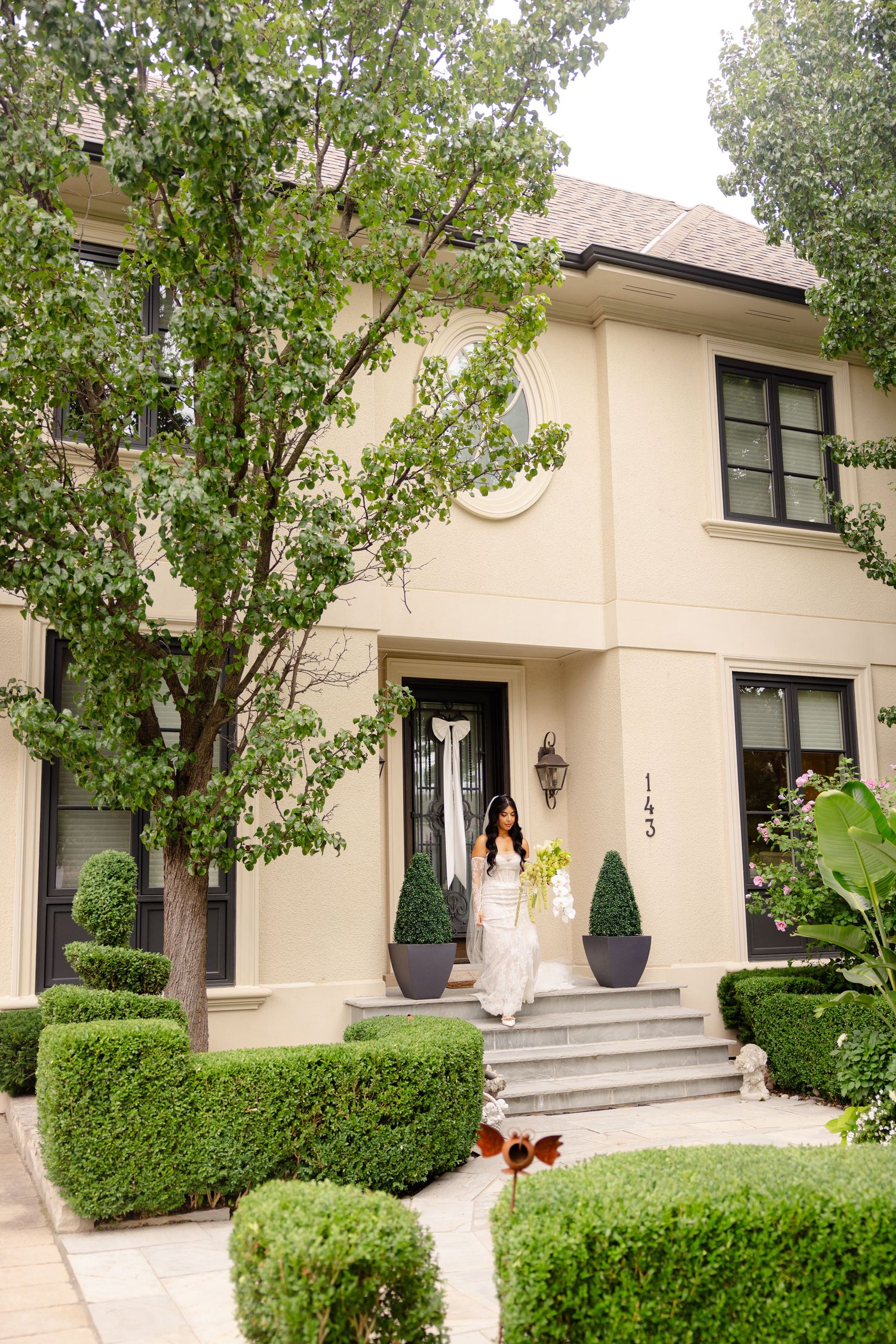 Landscaped entrance of a beige residential building with symmetrical bushes and mature trees.