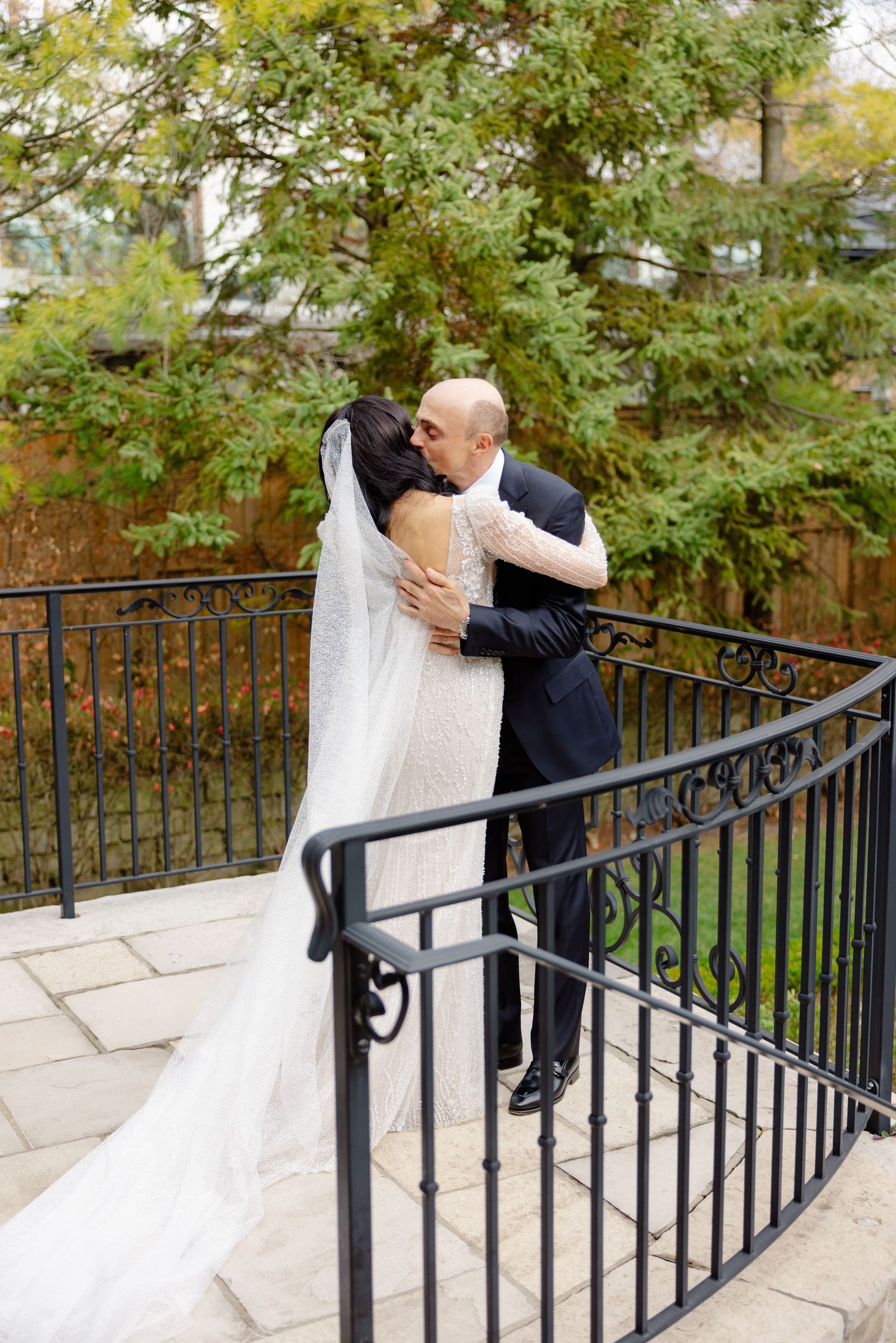 Newlyweds embrace on a garden terrace with wrought iron railings and autumn foliage in the background.