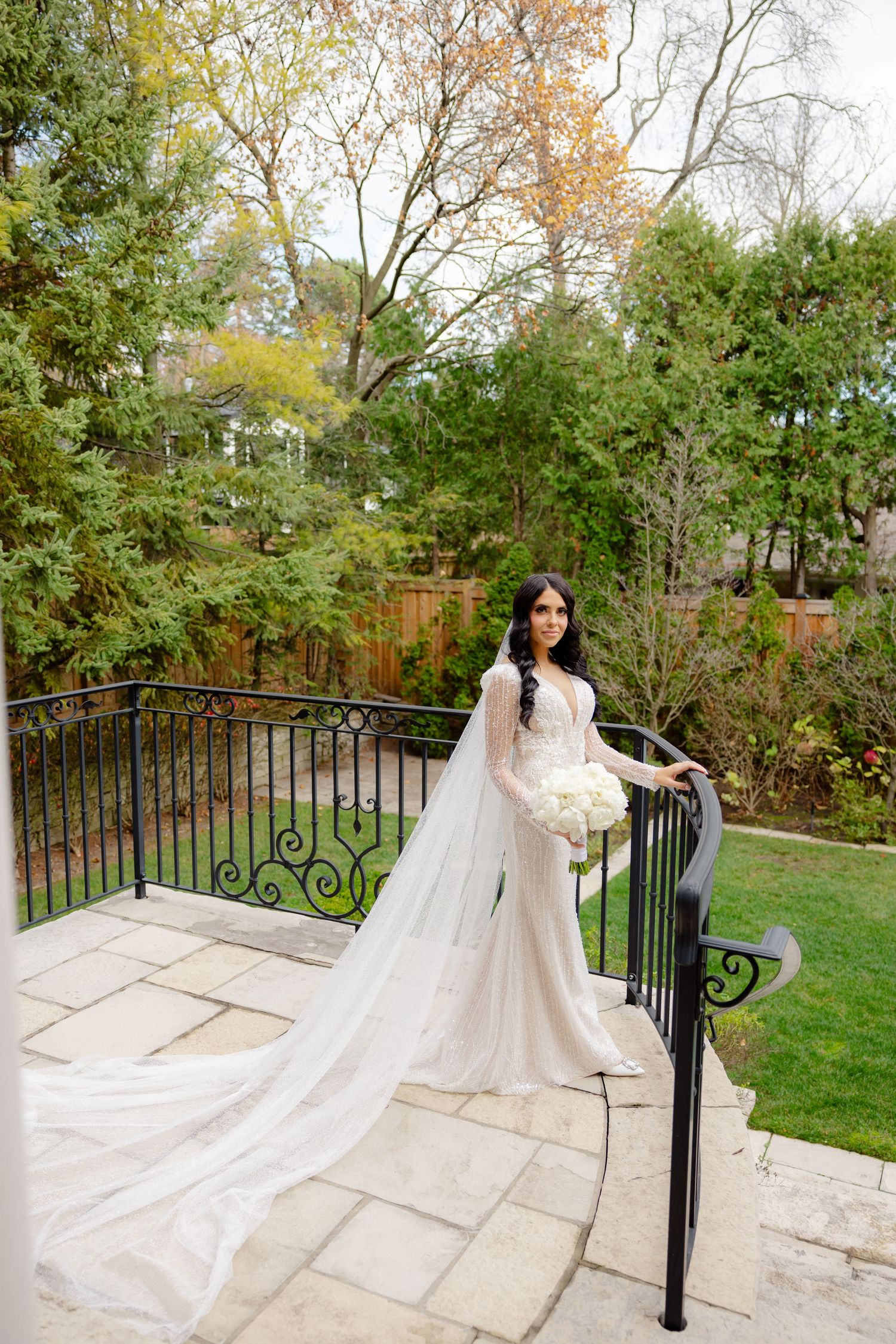 Bride in form-fitting dress and long veil poses by black iron railing with fall foliage in background.