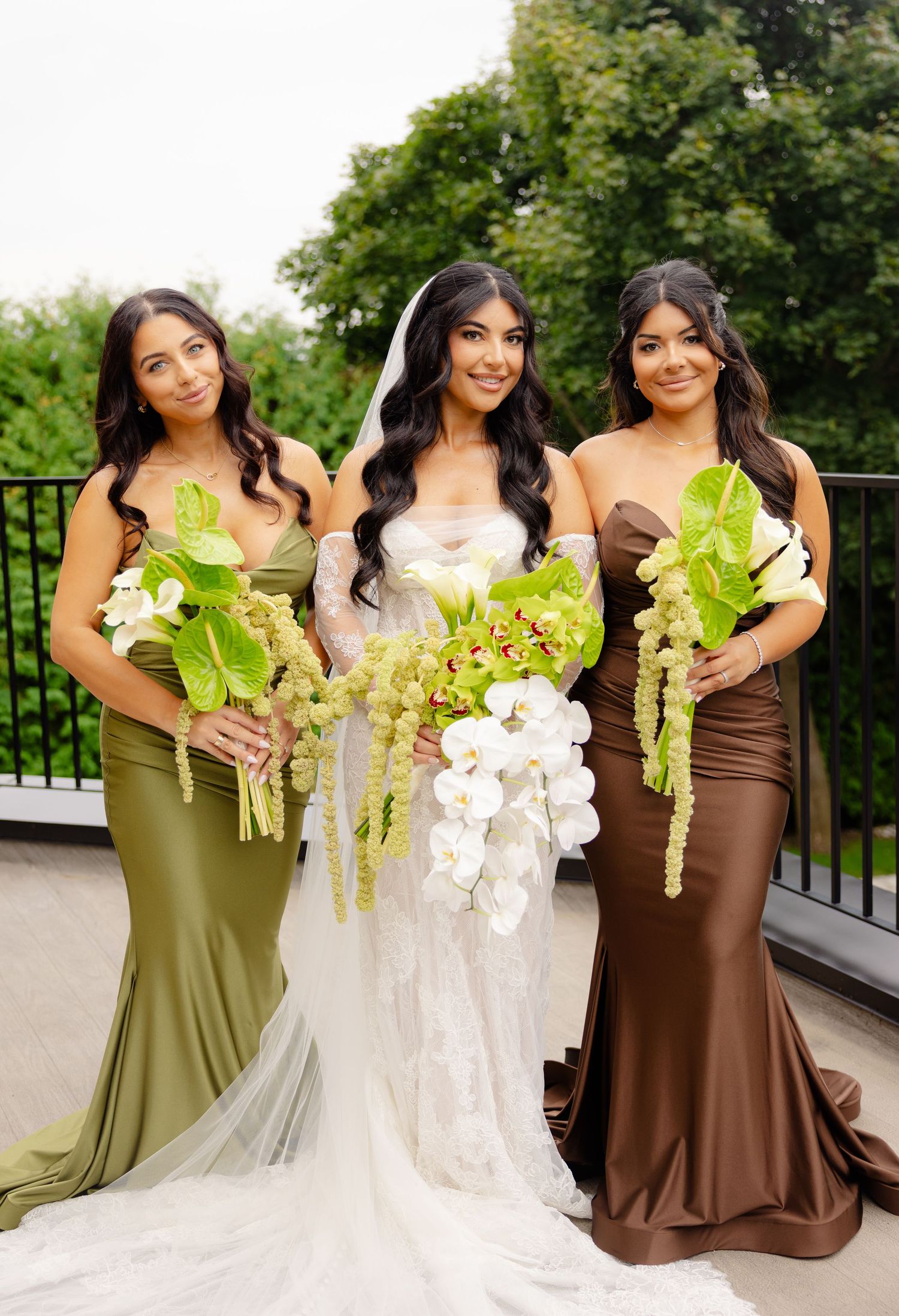 Three women in elegant dresses pose together at a wedding ceremony, holding beautiful floral bouquets.