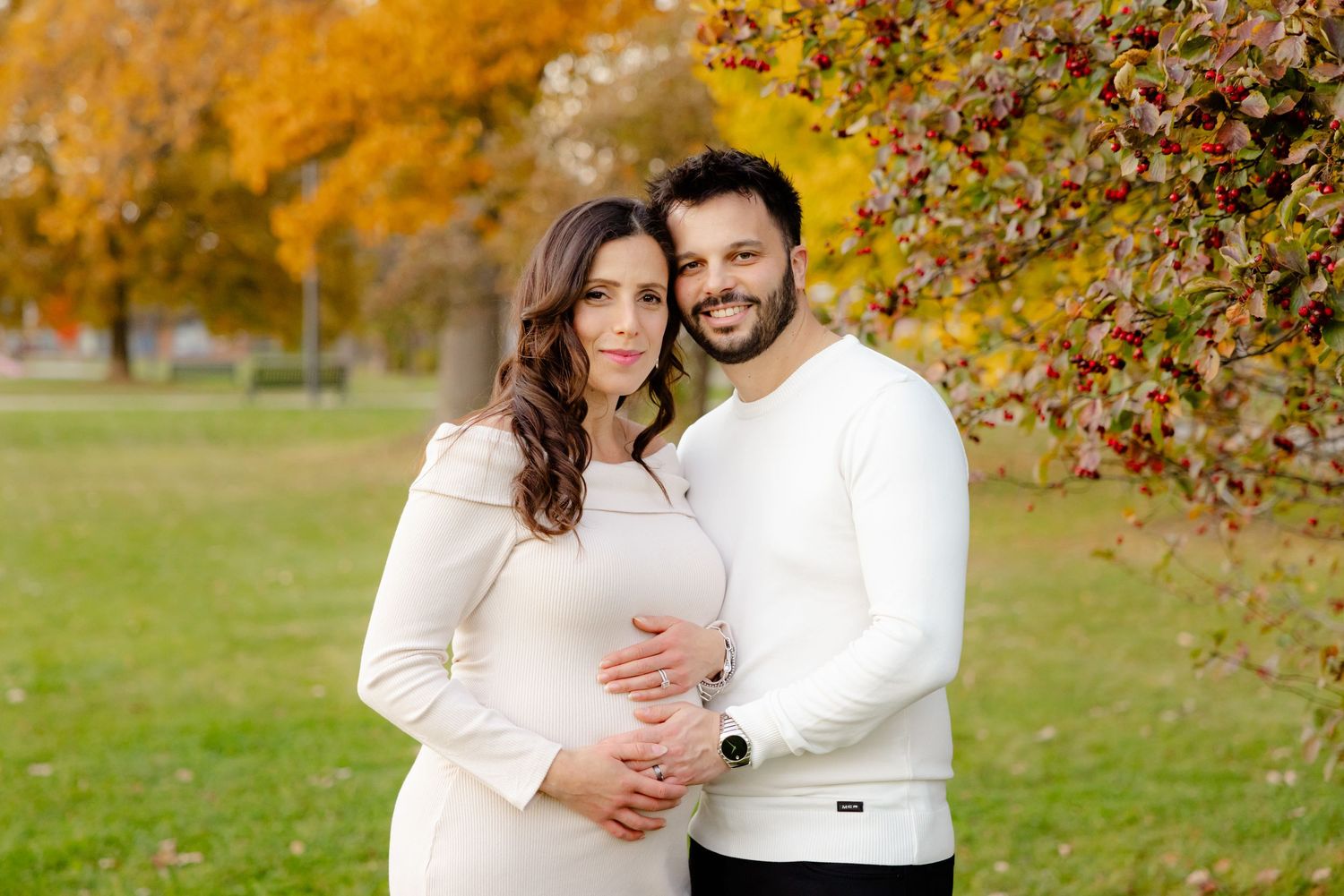 A couple in white sweaters embraces in an autumn park with colorful foliage in the background.