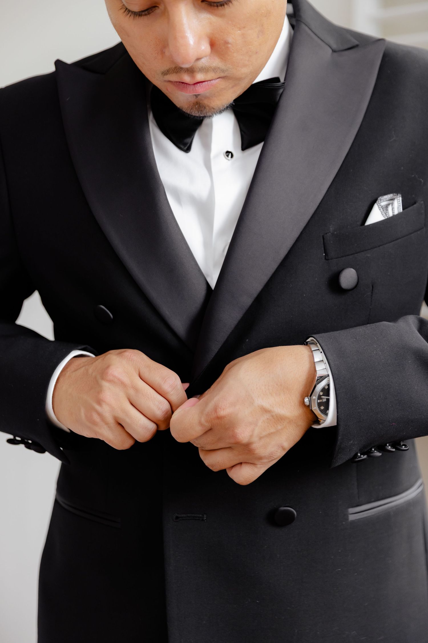 Close-up view of someone adjusting a black tuxedo jacket with white pocket square and bow tie, wearing a silver watch.