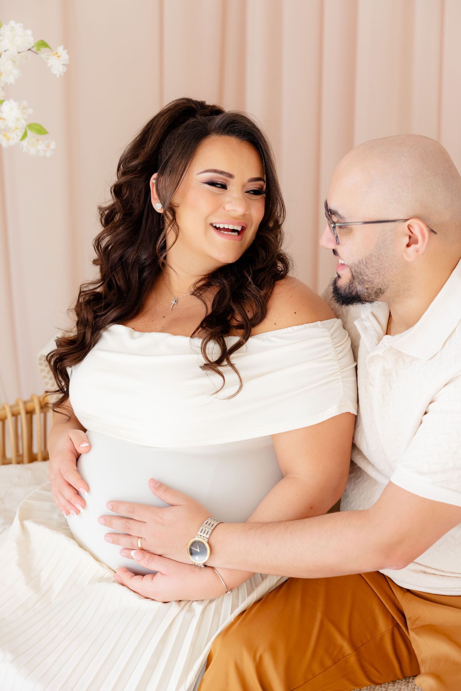 Happy couple in white outfits sharing an intimate moment while sitting together in a light-filled room with floral decor.