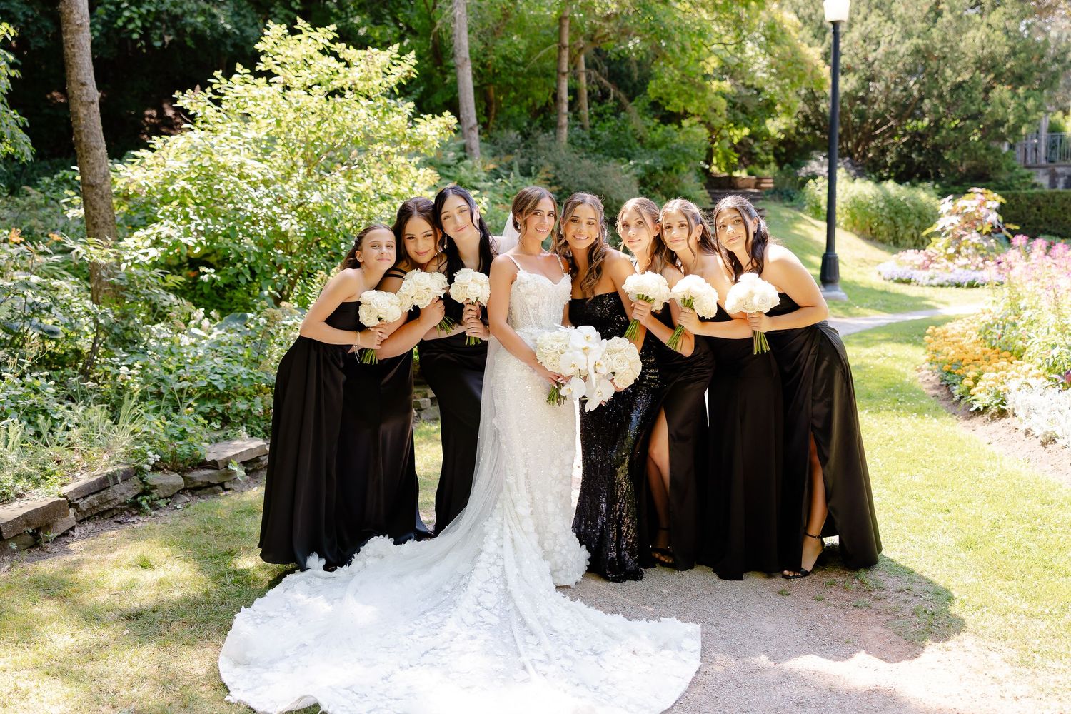 A bridal party in black bridesmaid dresses and white wedding gown pose together on a garden path in natural sunlight.