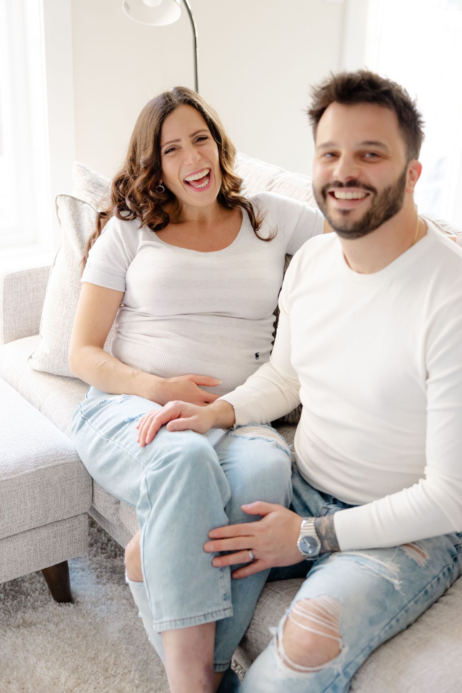 Smiling couple in white shirts sitting together on a couch, with the woman appearing to be pregnant.