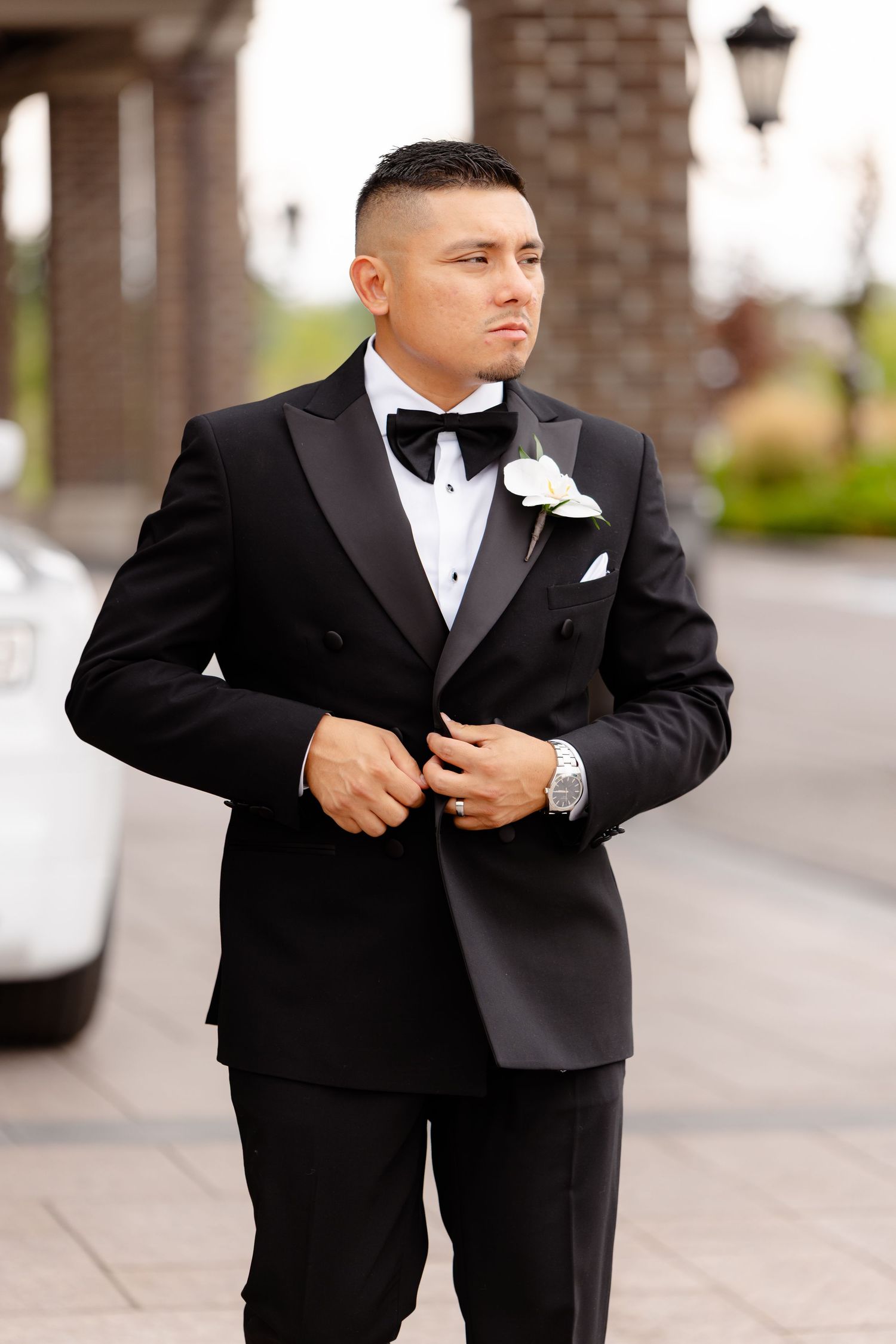 A groom in a classic black tuxedo with bowtie and white boutonniere poses outside an elegant brick building.