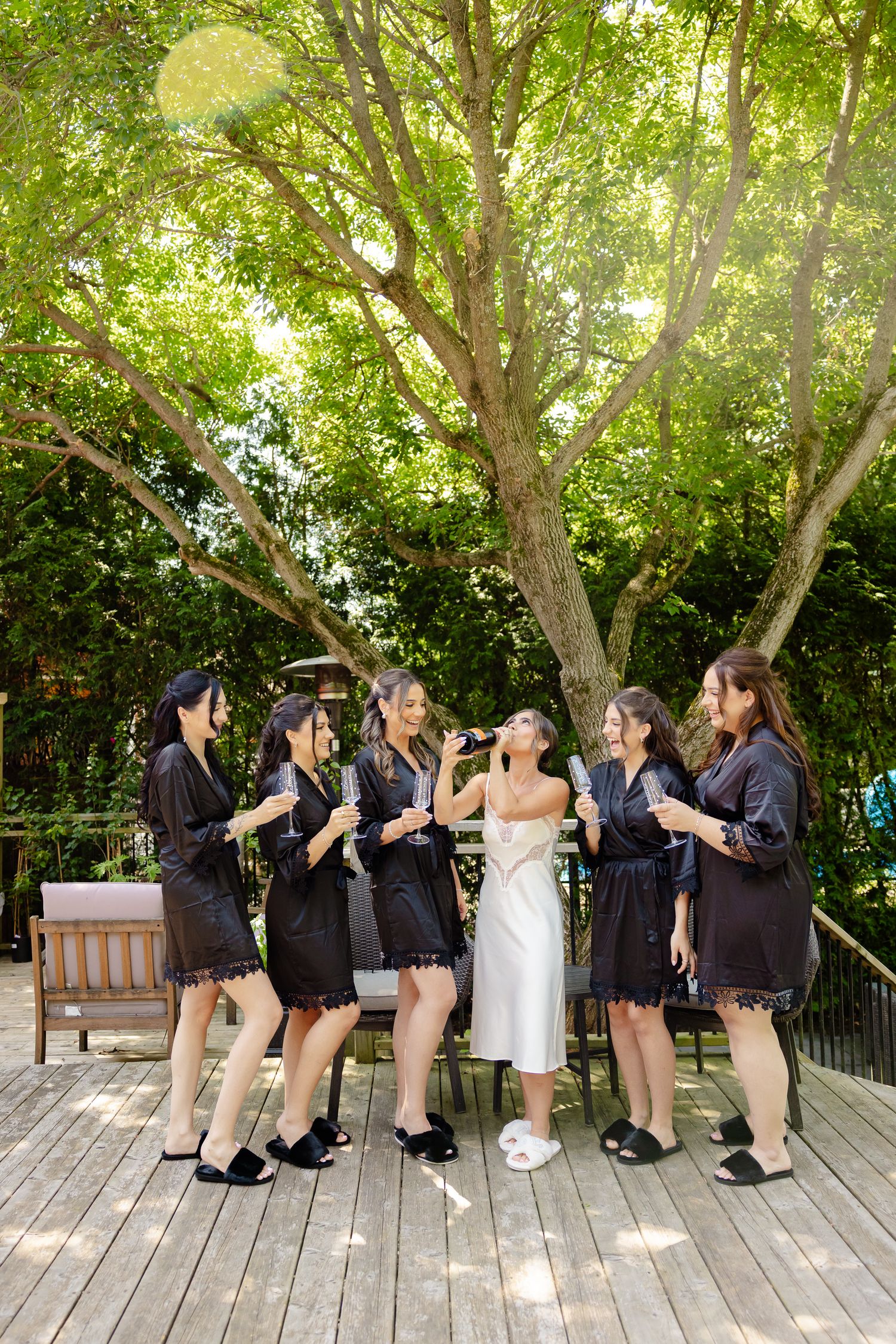 A wedding party wearing matching black robes and a white dress gather on a wooden deck surrounded by lush green trees.
