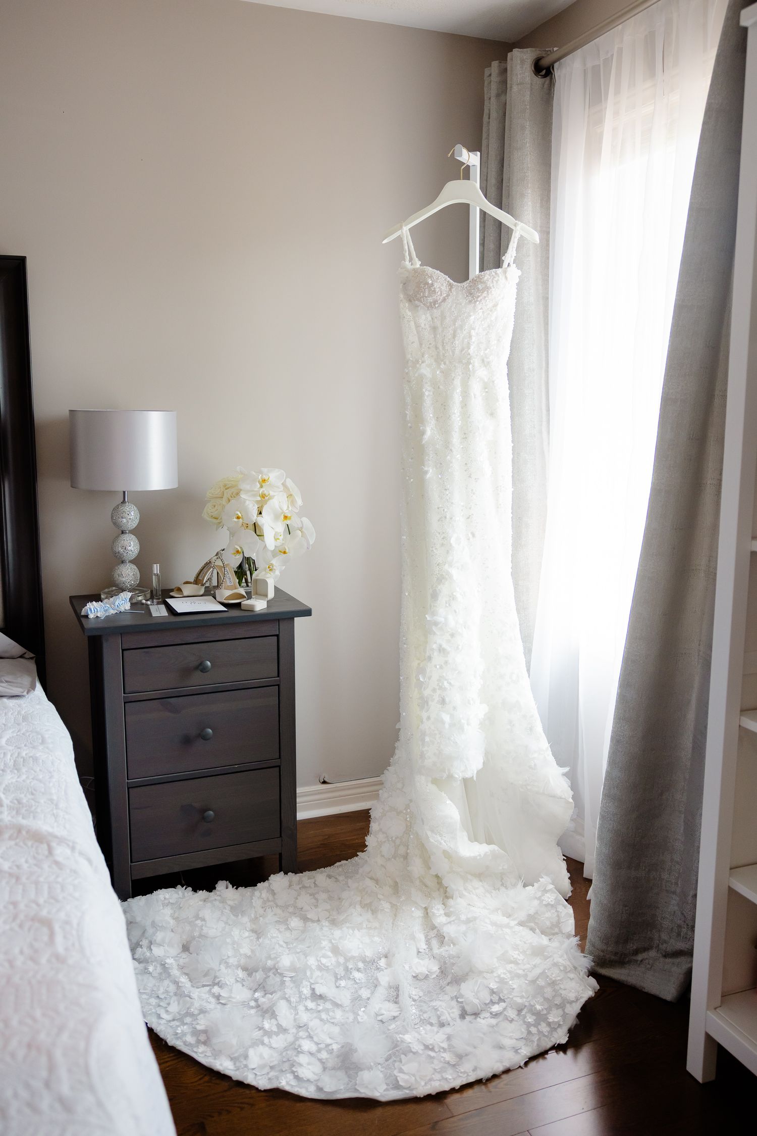 A row of elegant white wedding dresses displayed in a bedroom with dark furniture and sheer curtains.
