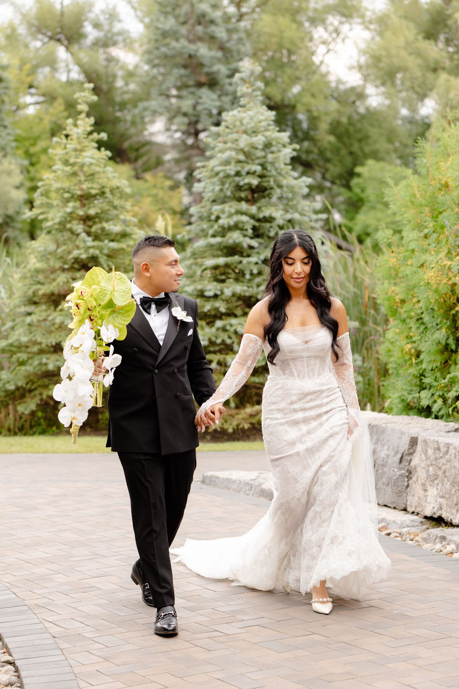 A wedding couple strolls along a stone path surrounded by lush greenery and trees in a garden setting.