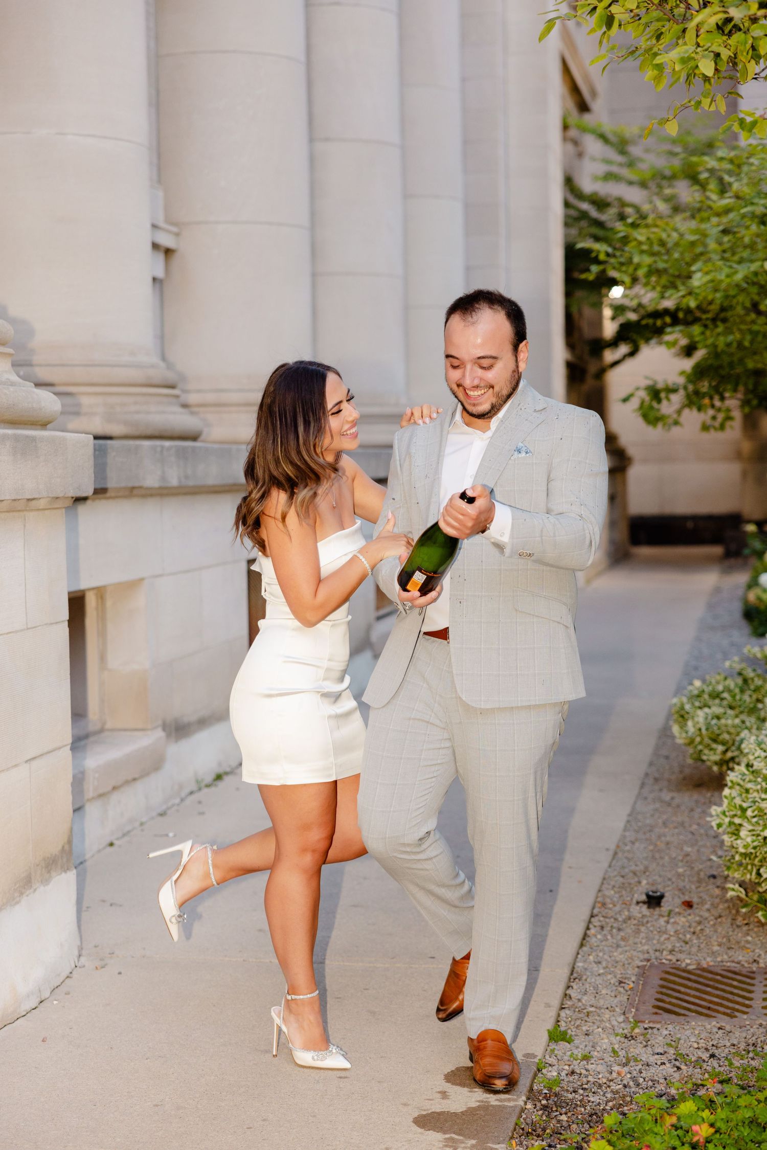 A couple jumps with excitement while holding a champagne bottle by architectural columns.