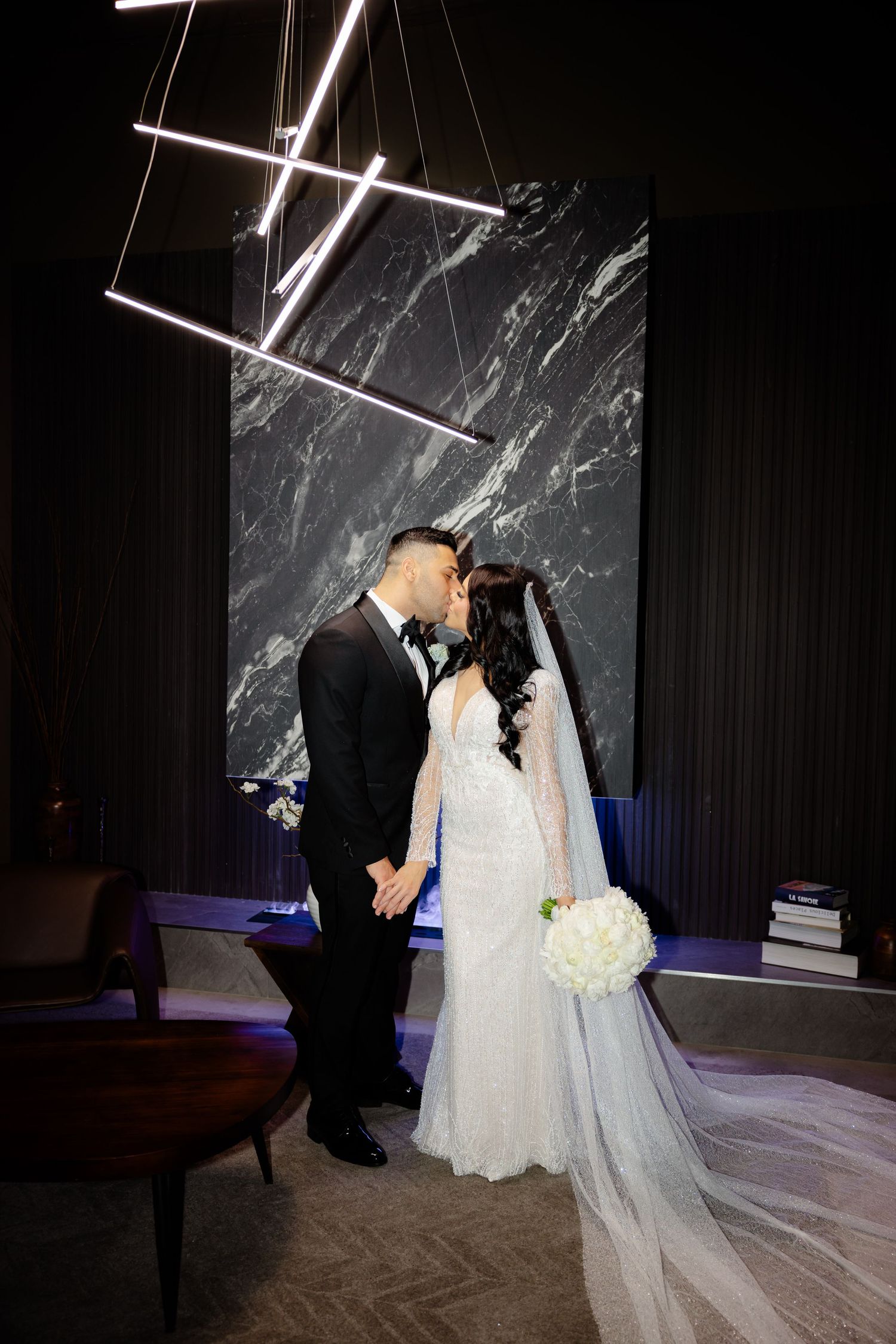 A romantic wedding moment against a marble wall backdrop with modern lighting fixtures and a white floral bouquet.