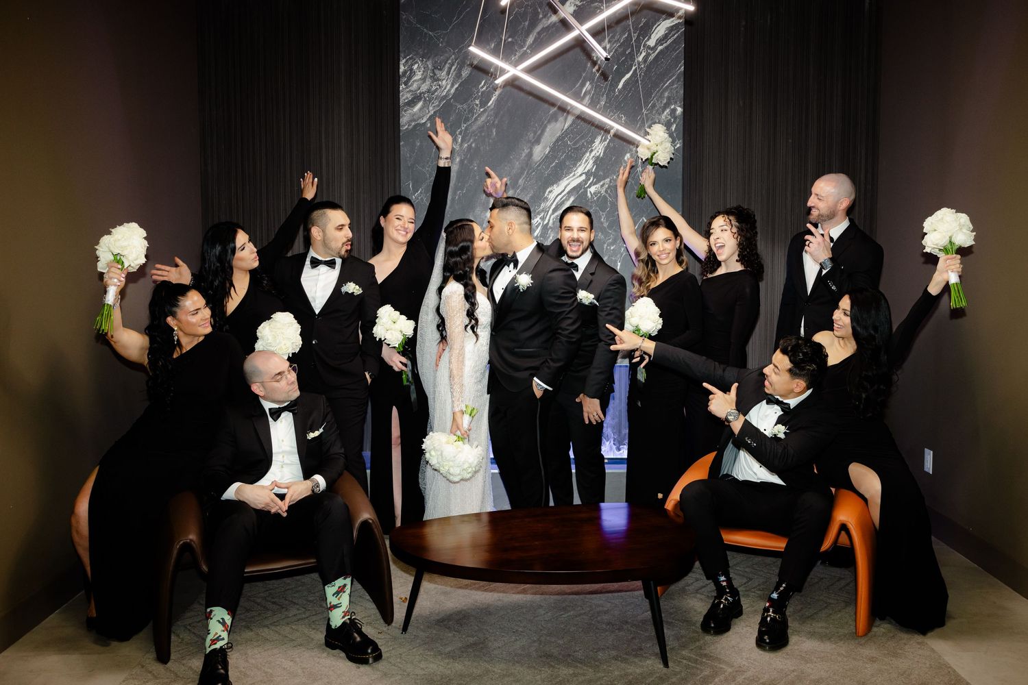 Wedding party celebrates energetically with raised arms against marble backdrop with modern lighting.