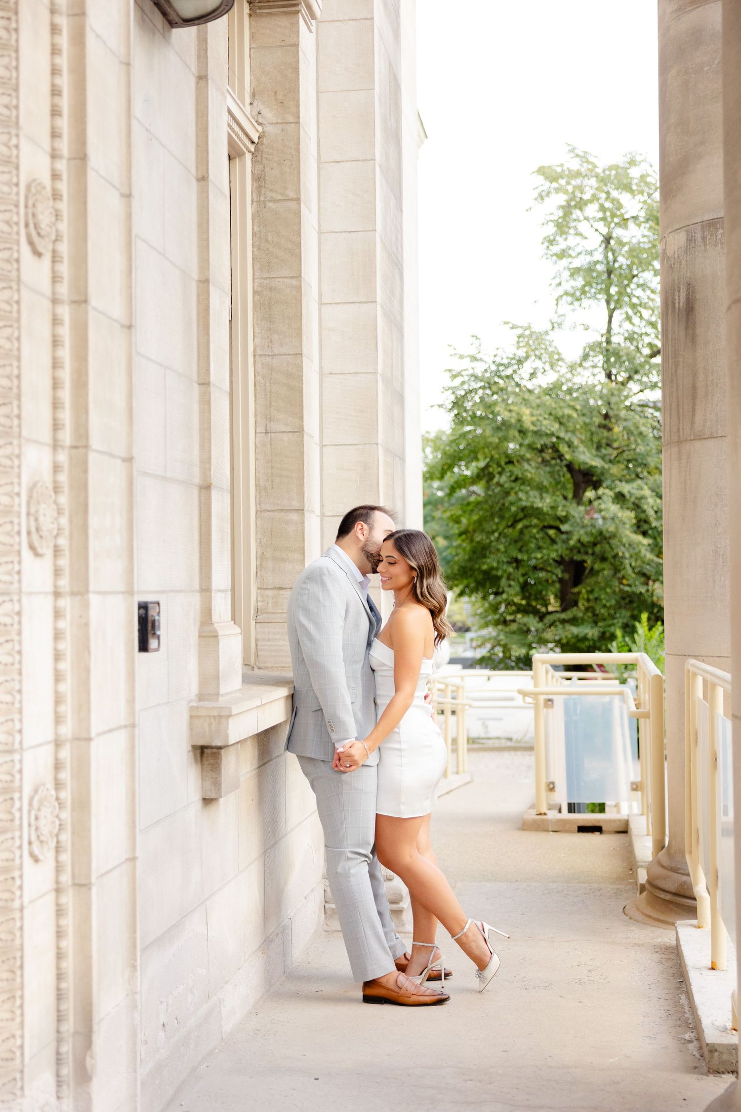 A romantic couple embraces against a stone column on an ornate balcony walkway.