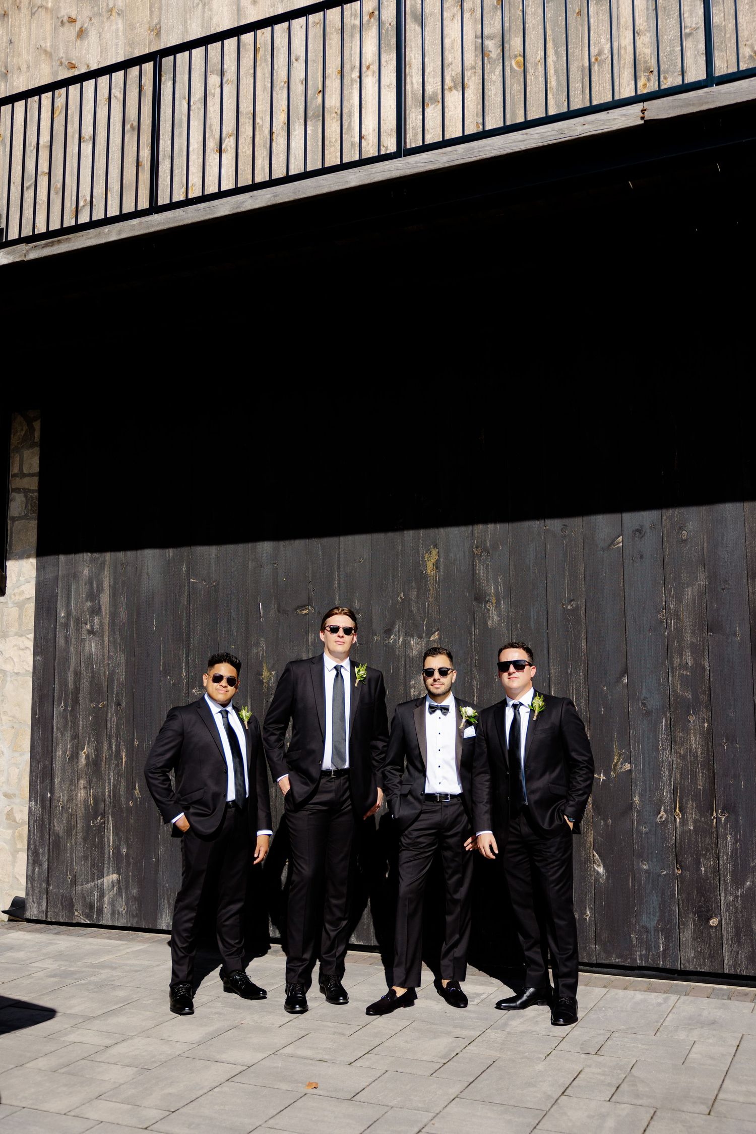 Groomsmen strike confident pose in matching dark suits and sunglasses against wooden backdrop.