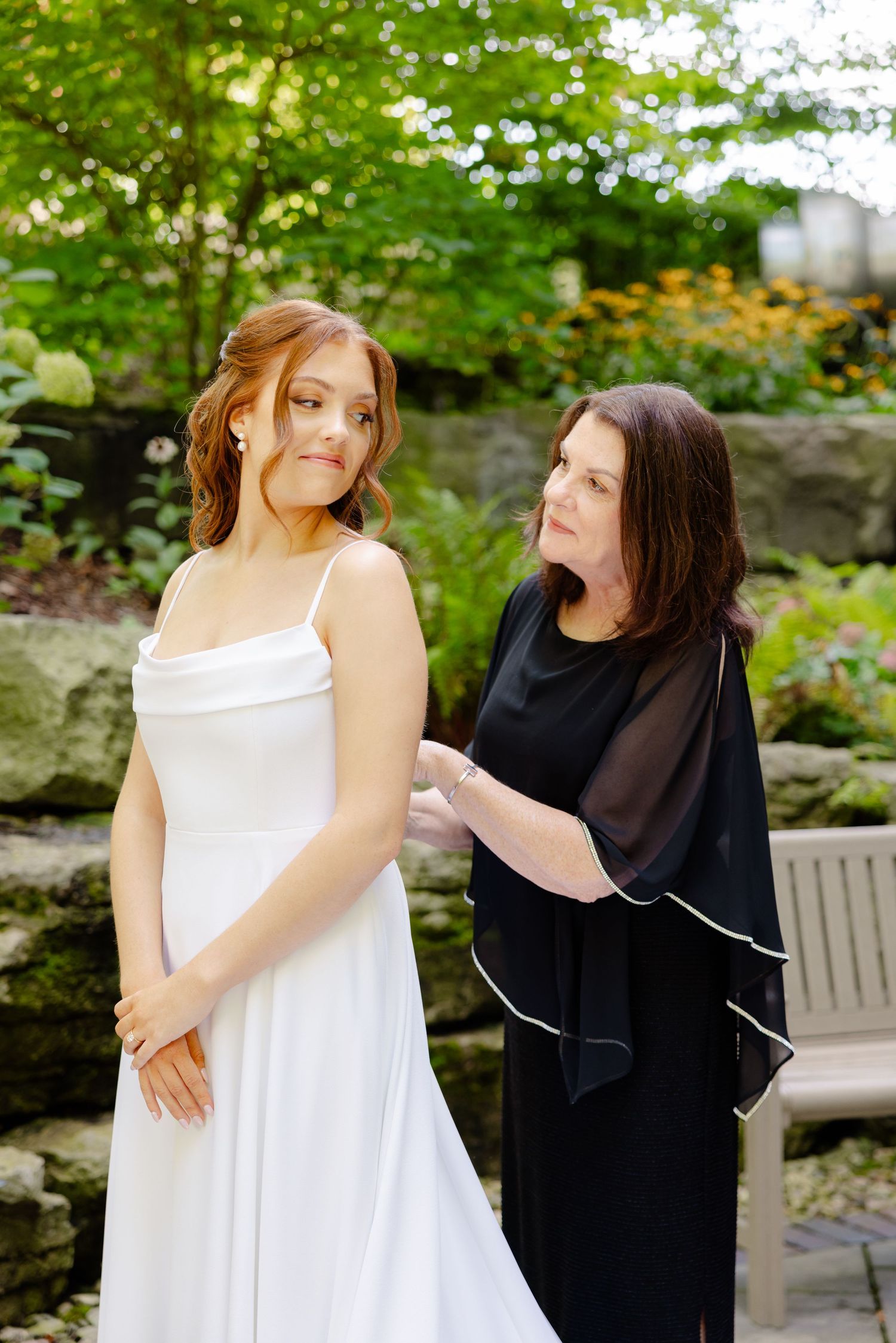 A woman in a white wedding gown gets help with her dress in a garden setting.