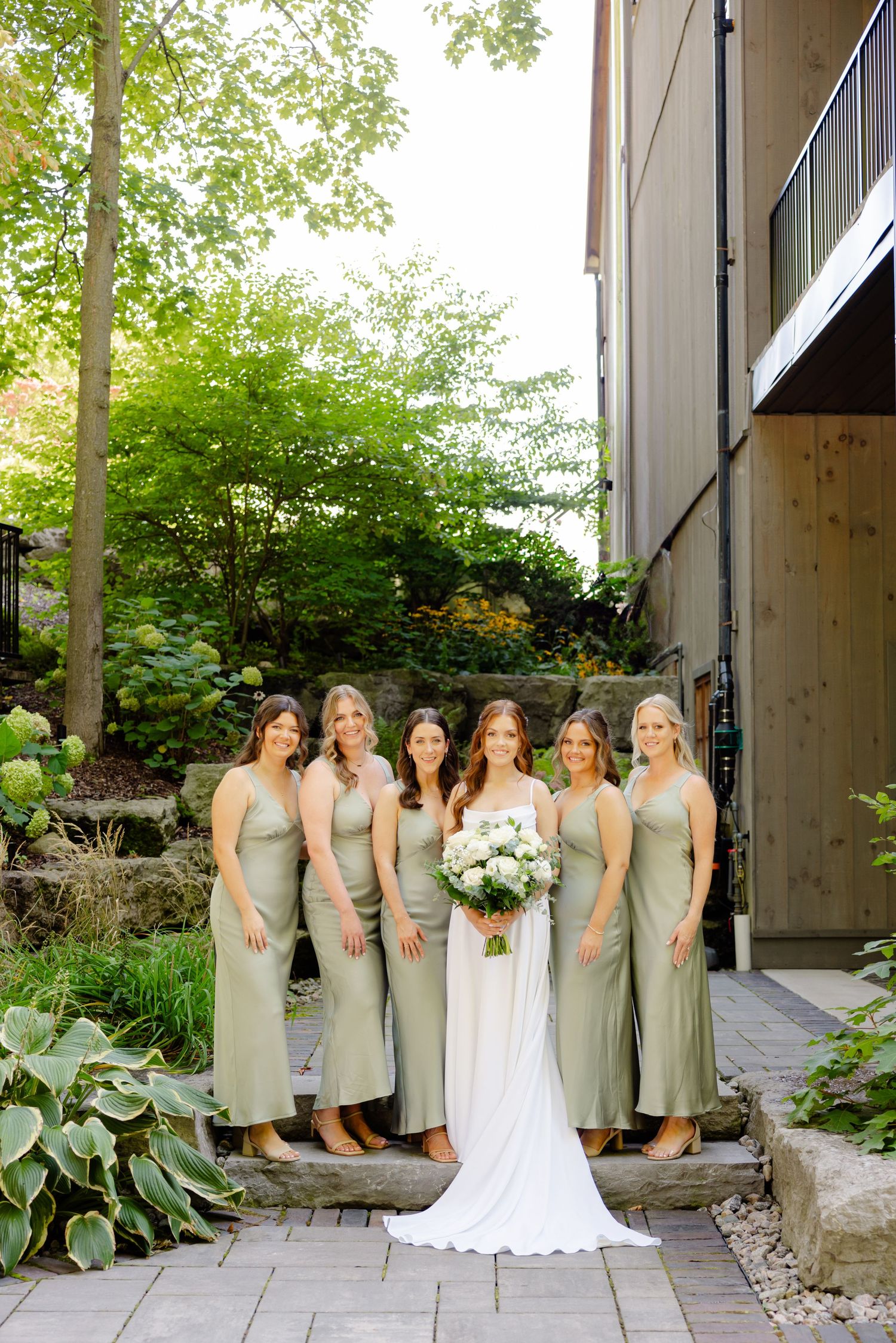 Bridal party poses outdoors wearing sage dresses against modern building and garden landscape.