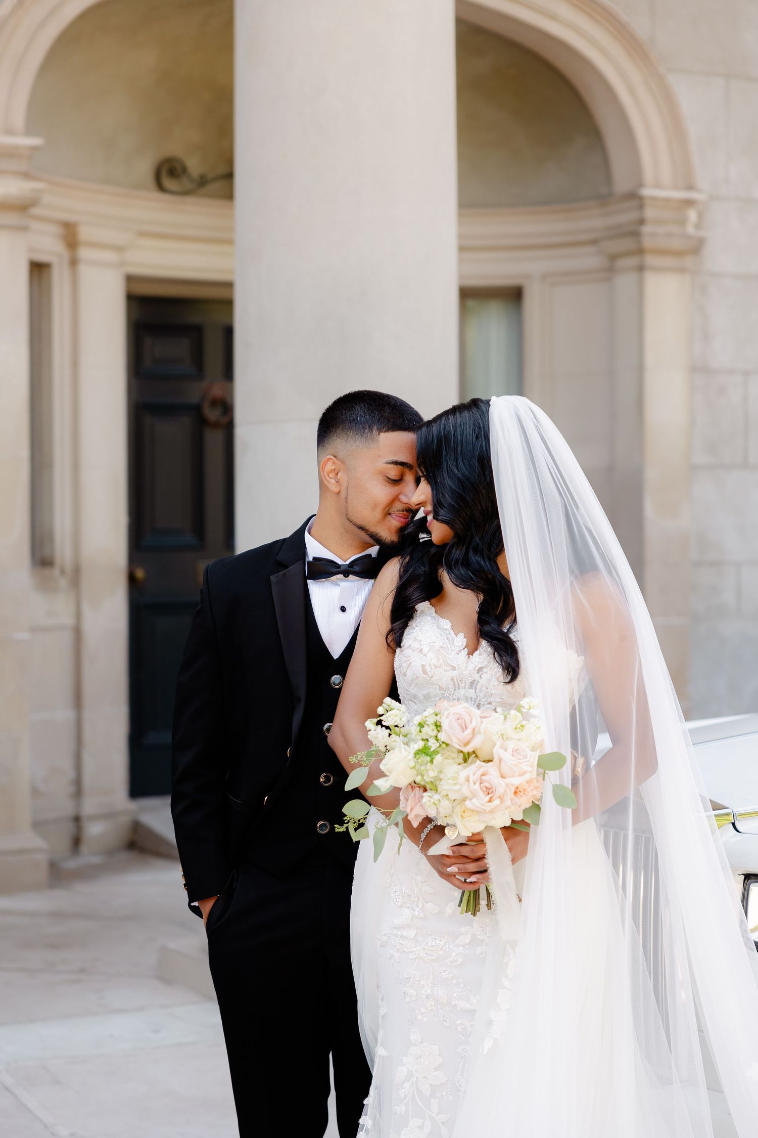 Intimate wedding portrait of couple by white pillars with bride holding delicate bouquet.