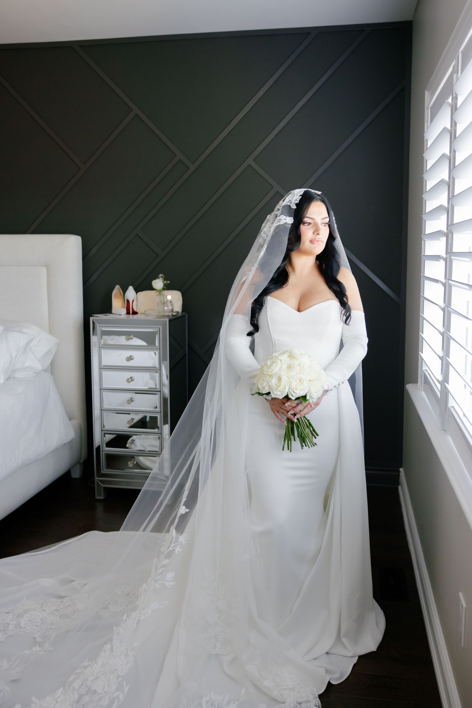 Stunning bride in long sleeve wedding dress with flowing veil poses in bedroom setting.