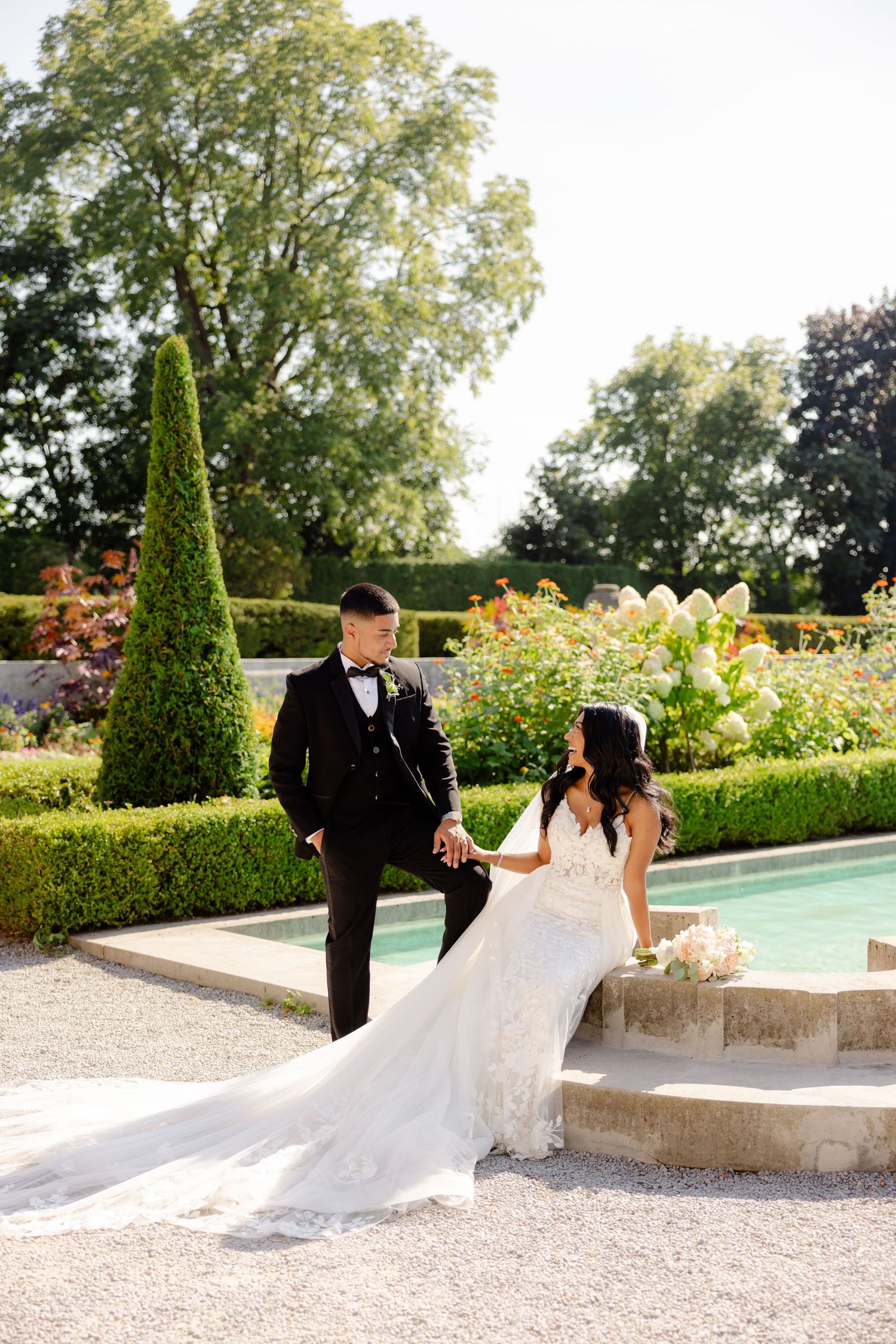 A couple poses together by stone steps in a formal garden with a reflecting pool and cypress trees.