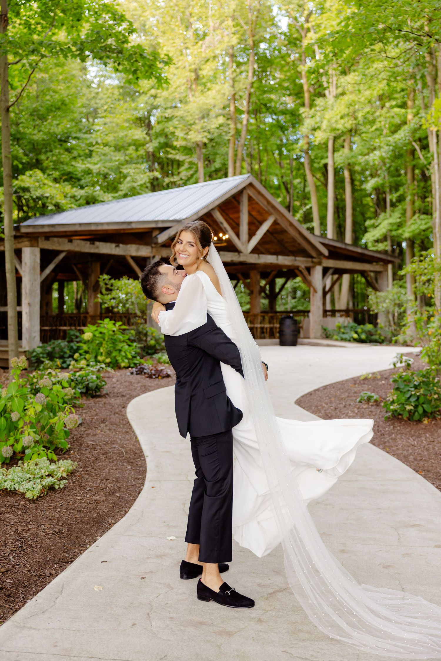 Groom lifts bride in an elegant spin on a curved pathway near a wooden pavilion.