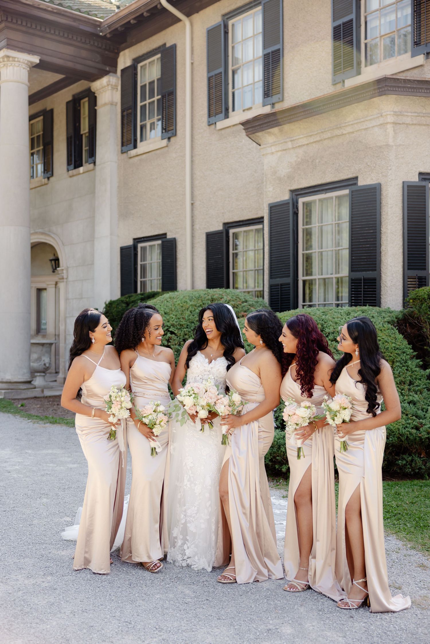 Bridesmaids in champagne gowns holding white bouquets pose outdoors at elegant venue.