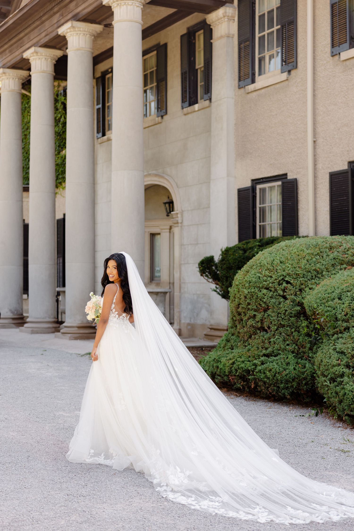 A radiant bride with long cathedral veil poses in front of a grand colonial-style building.