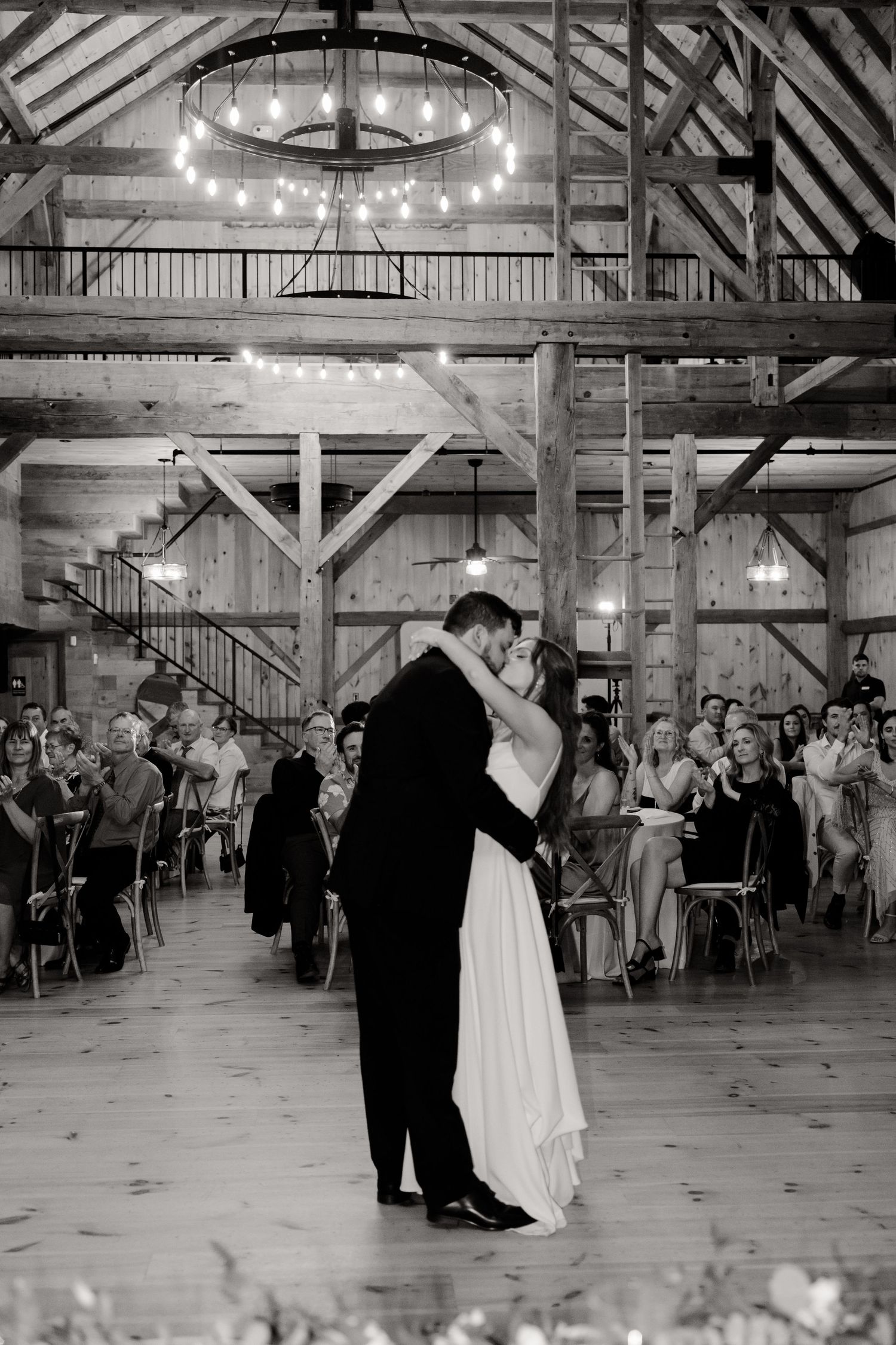 Black and white photo of couple dancing in barn venue with exposed beams and chandeliers.