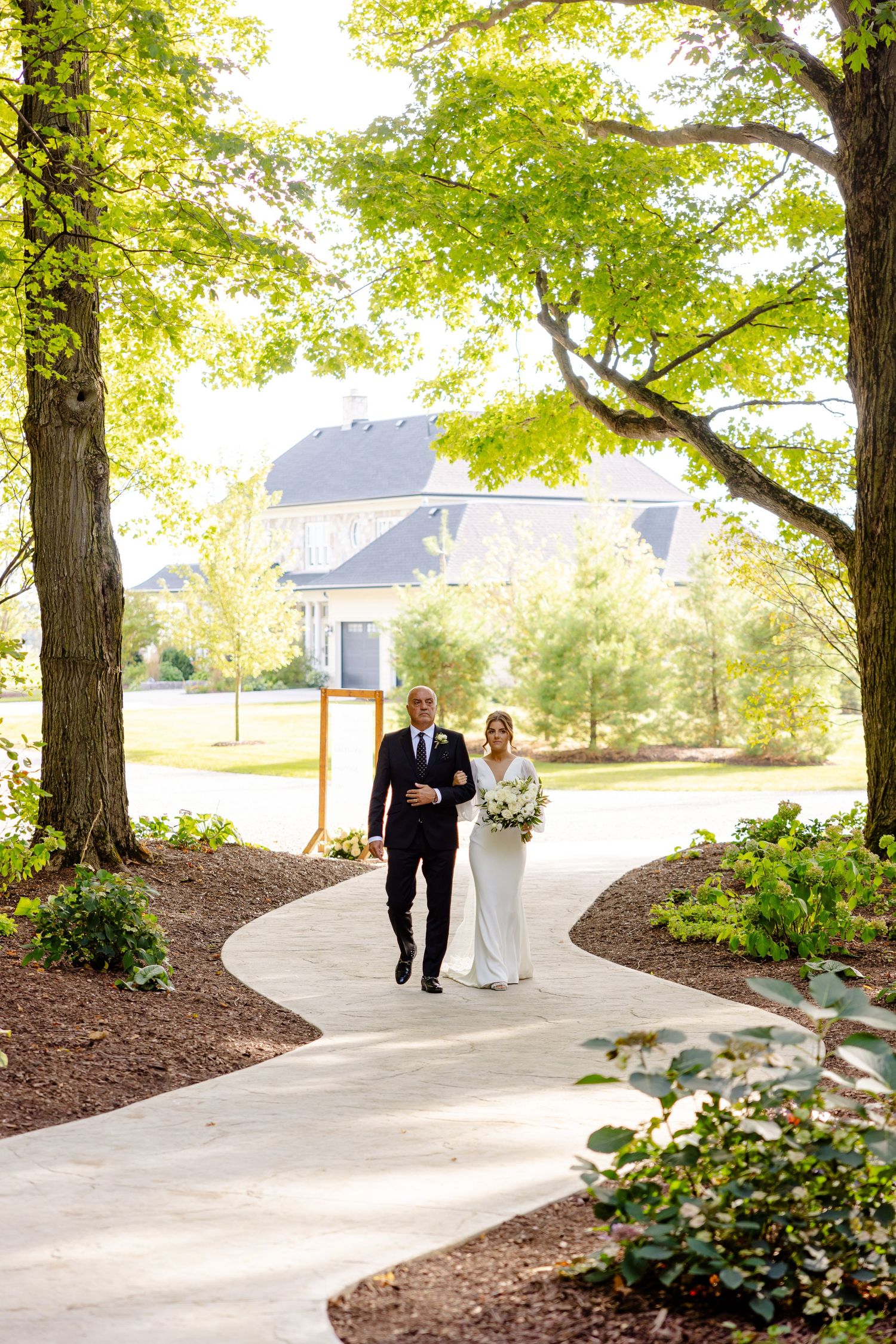 A couple walks down a winding path surrounded by lush green trees on their wedding day.