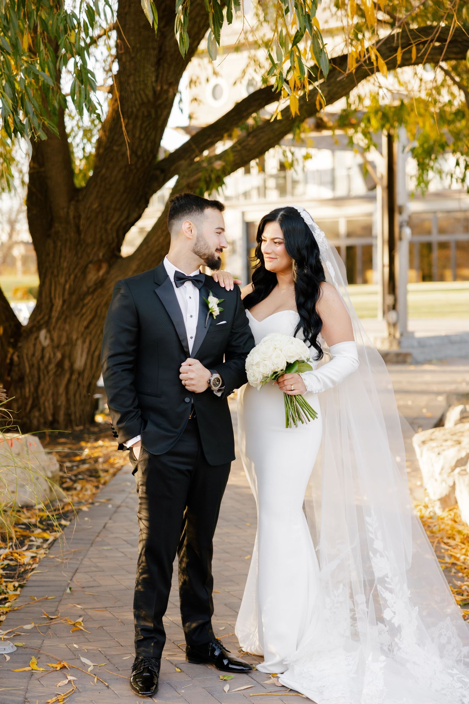 Elegant wedding couple shares intimate moment under willow tree branches on stone path.
