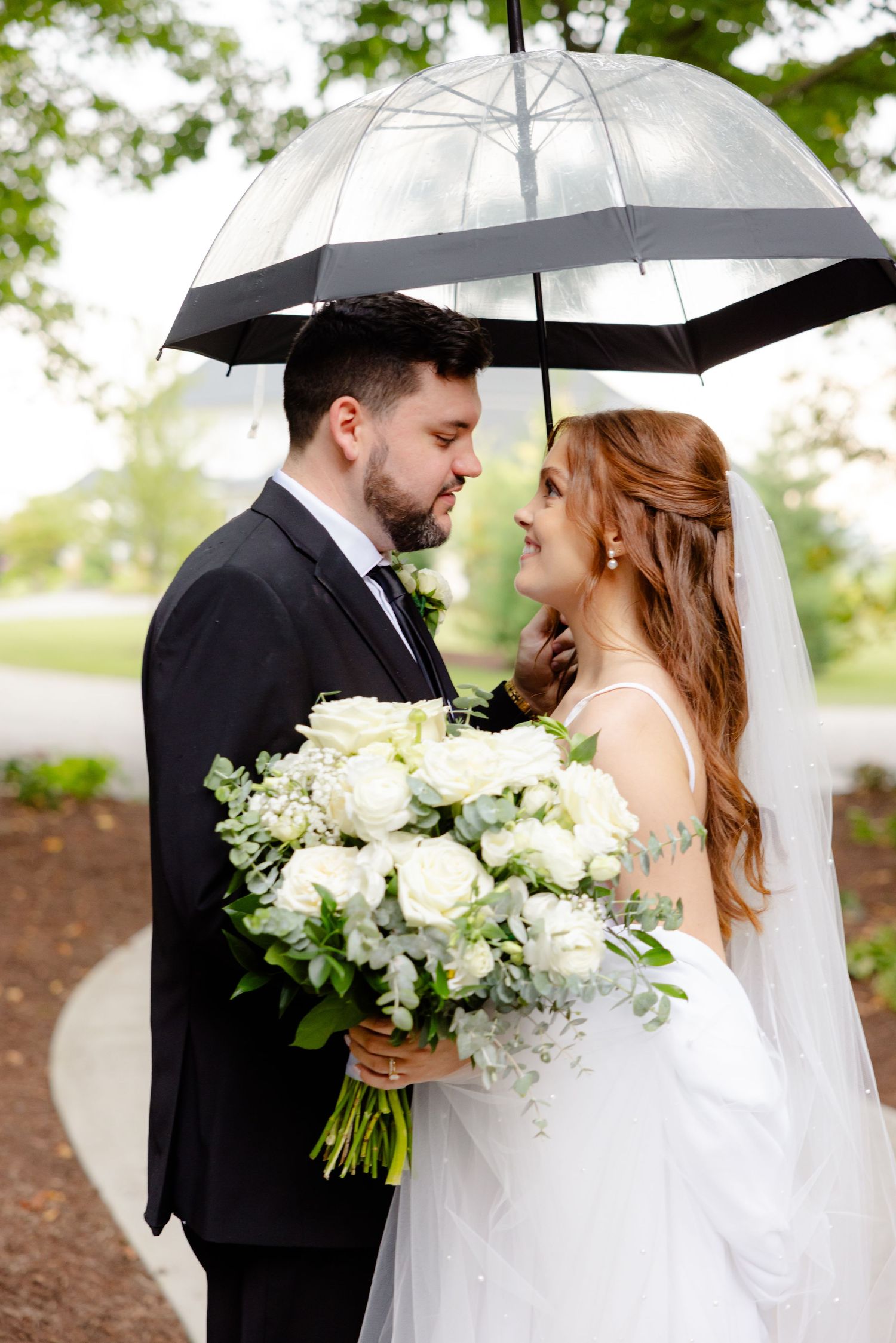 A wedding couple shares an intimate moment under a clear umbrella holding a white floral bouquet in an outdoor setting.