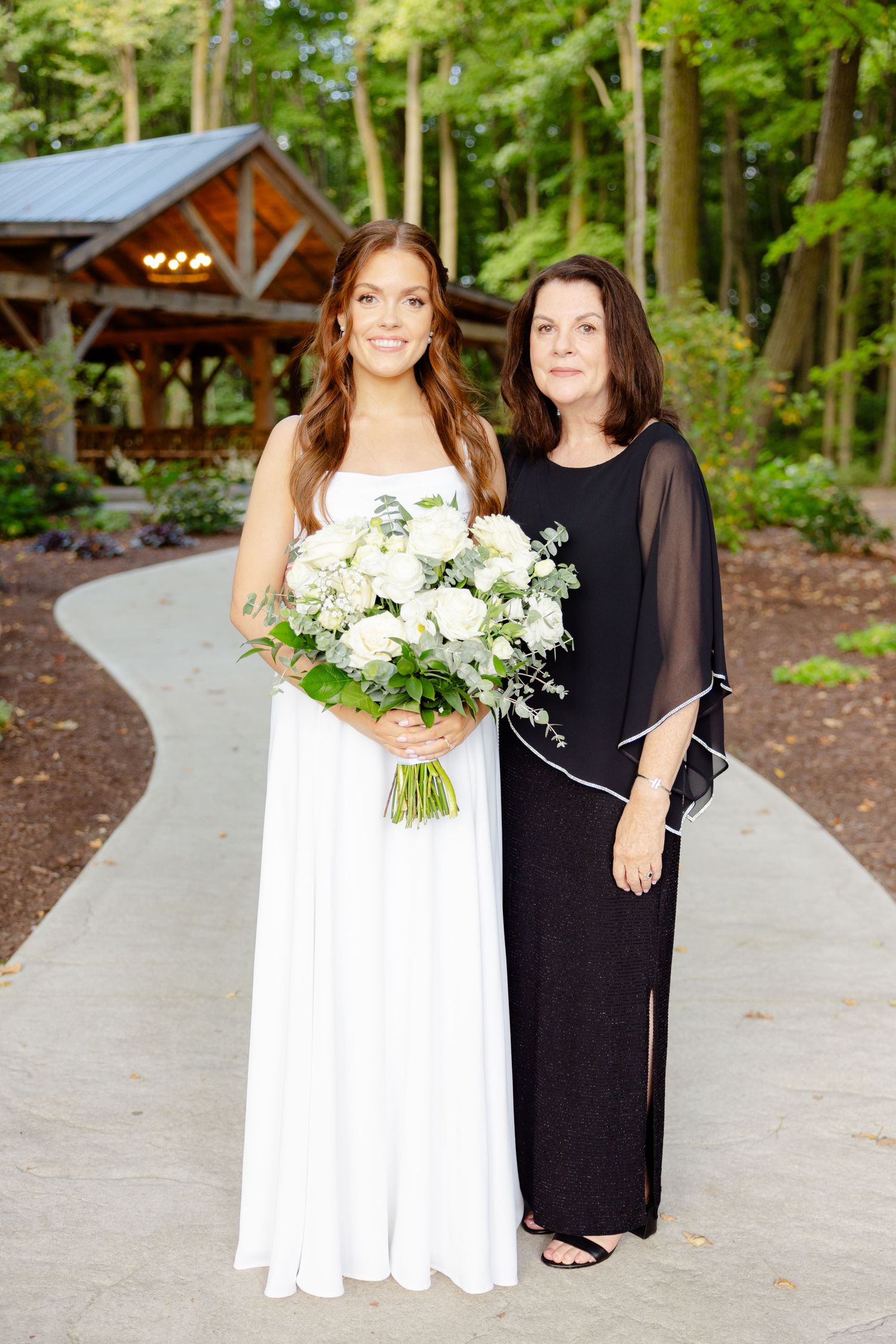Wedding portrait taken on a concrete path with rustic wooden pavilion and forest backdrop.