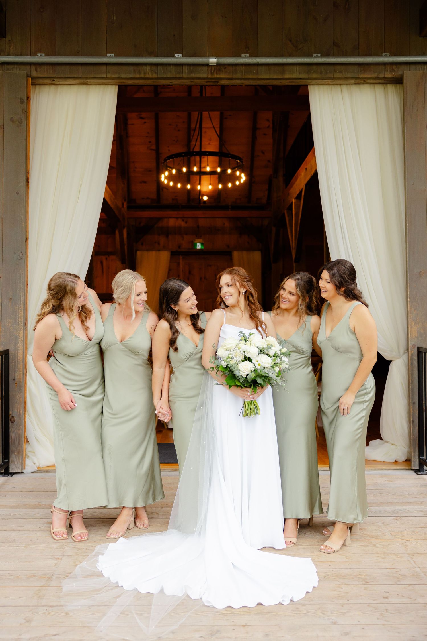 Wedding party sharing candid moment at barn entrance with white curtains and chandelier.