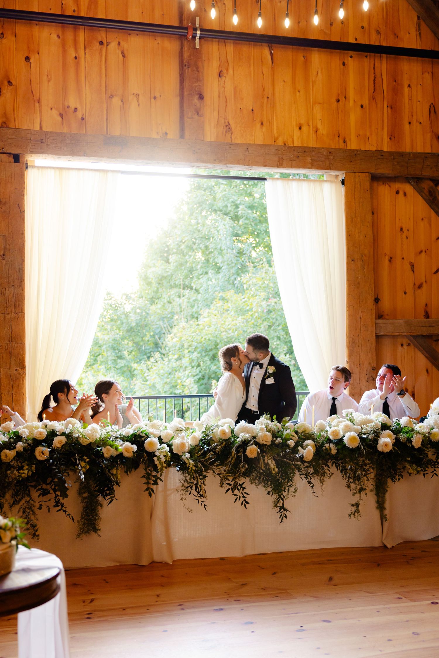 Wedding head table decorated with white floral garland in a sunlit rustic barn venue.