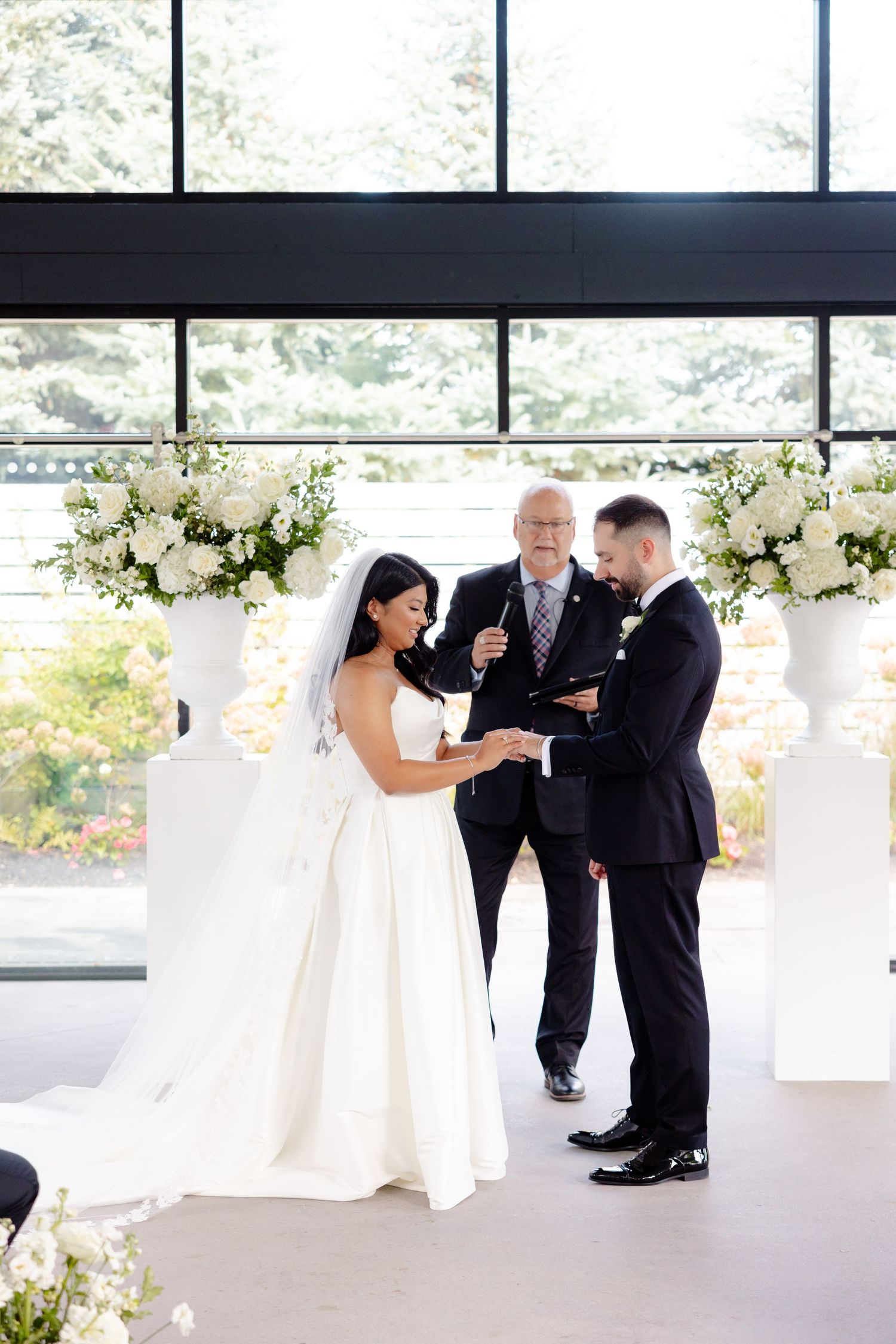 A wedding ceremony exchange in a bright venue decorated with elegant white floral arrangements on pedestals.