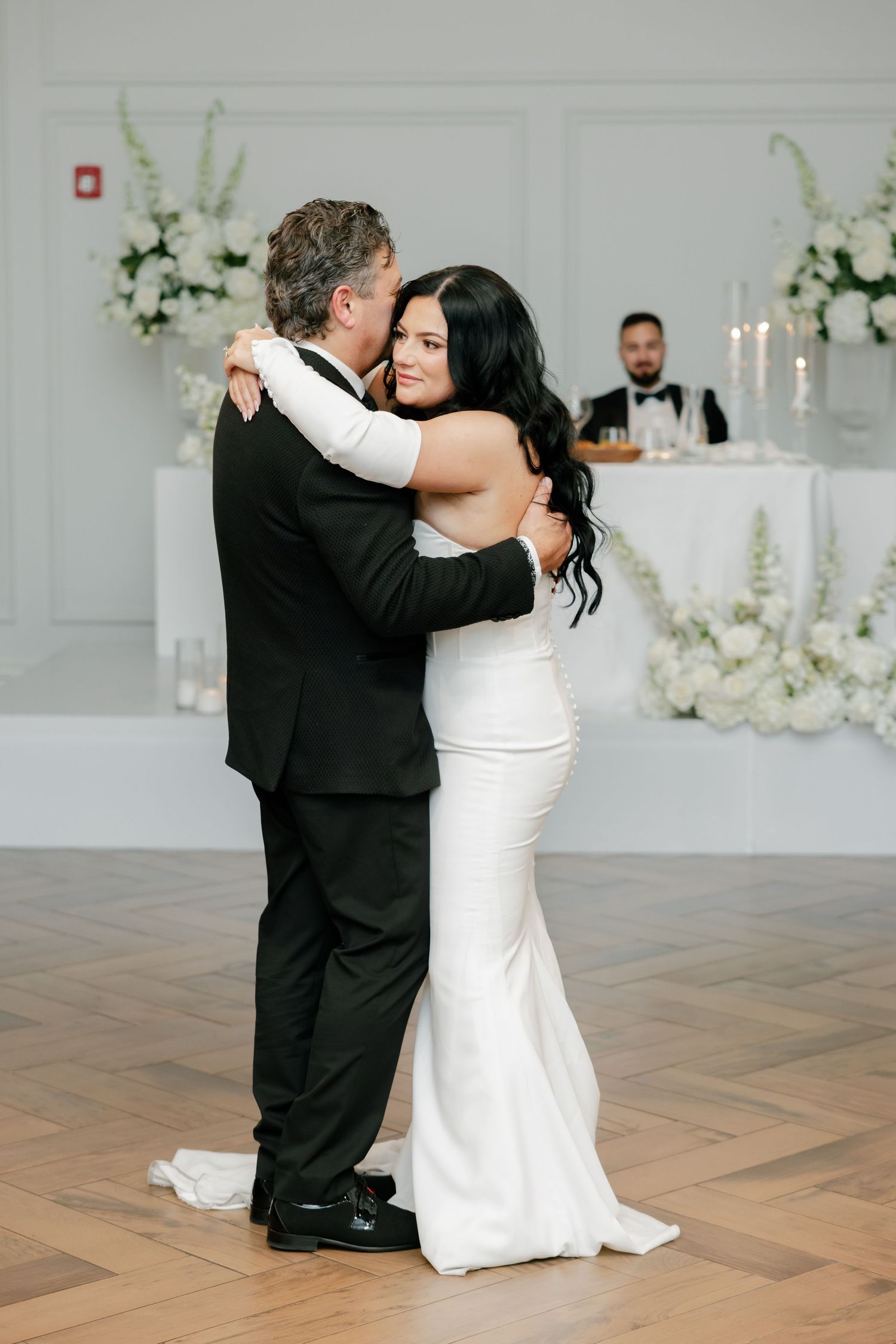 A tender moment between newlyweds dancing in an elegant white room decorated with floral arrangements.