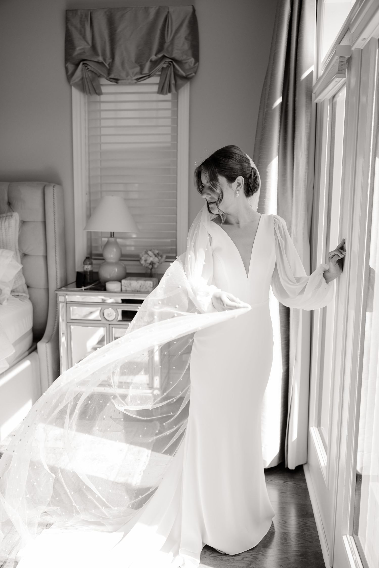 Black and white photo of a wedding dress with dramatic sleeves and train by a window.