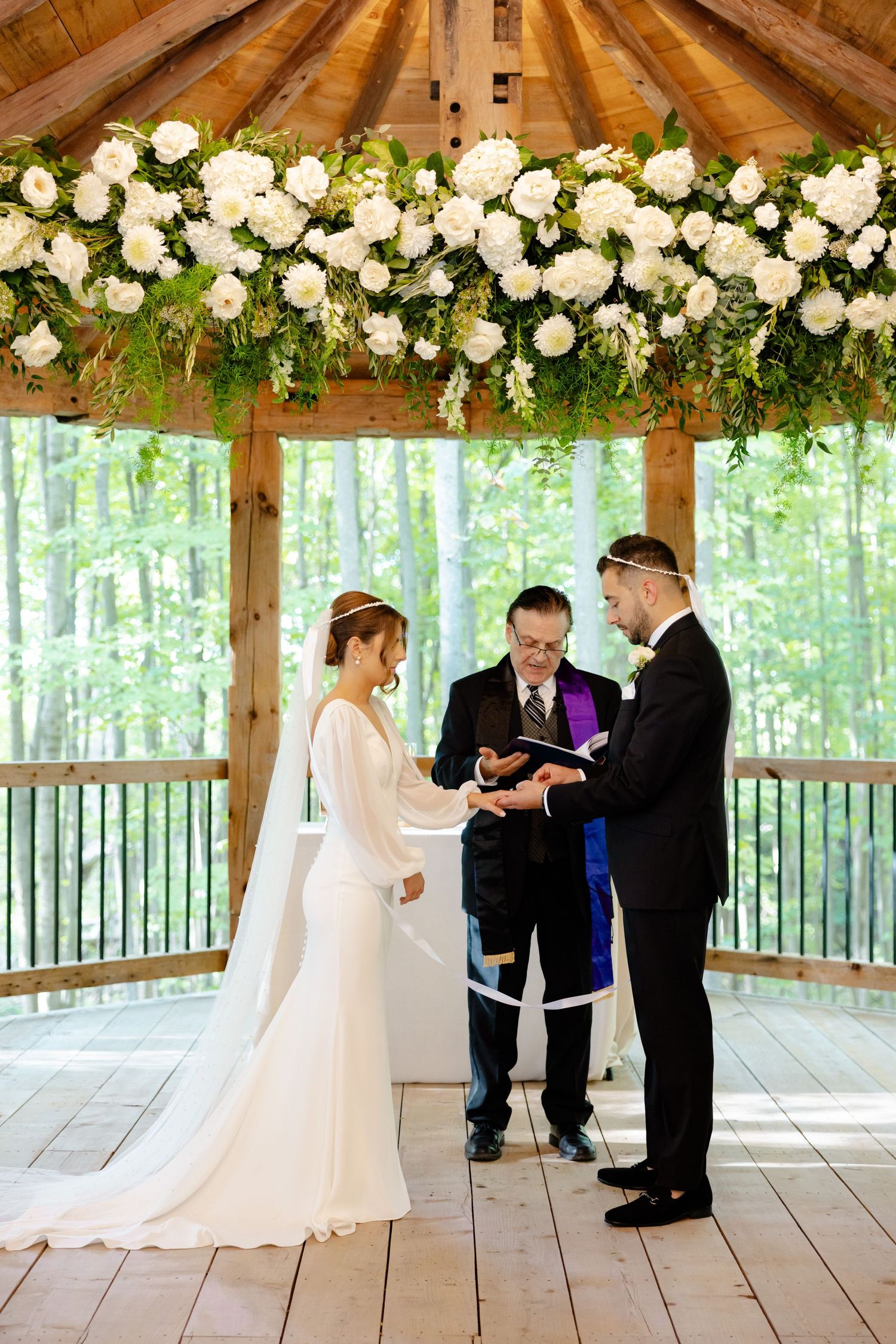 Wedding ceremony with white floral arch in rustic wooden pavilion overlooking wooded scenery.
