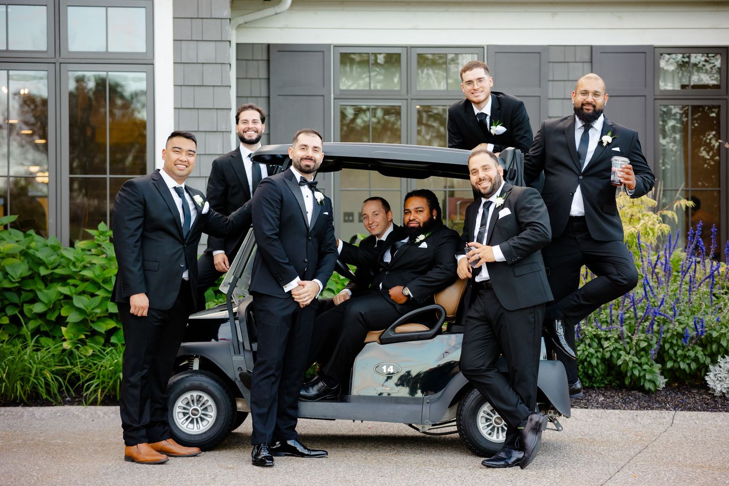 Wedding party poses playfully with golf cart outside venue surrounded by landscaping.