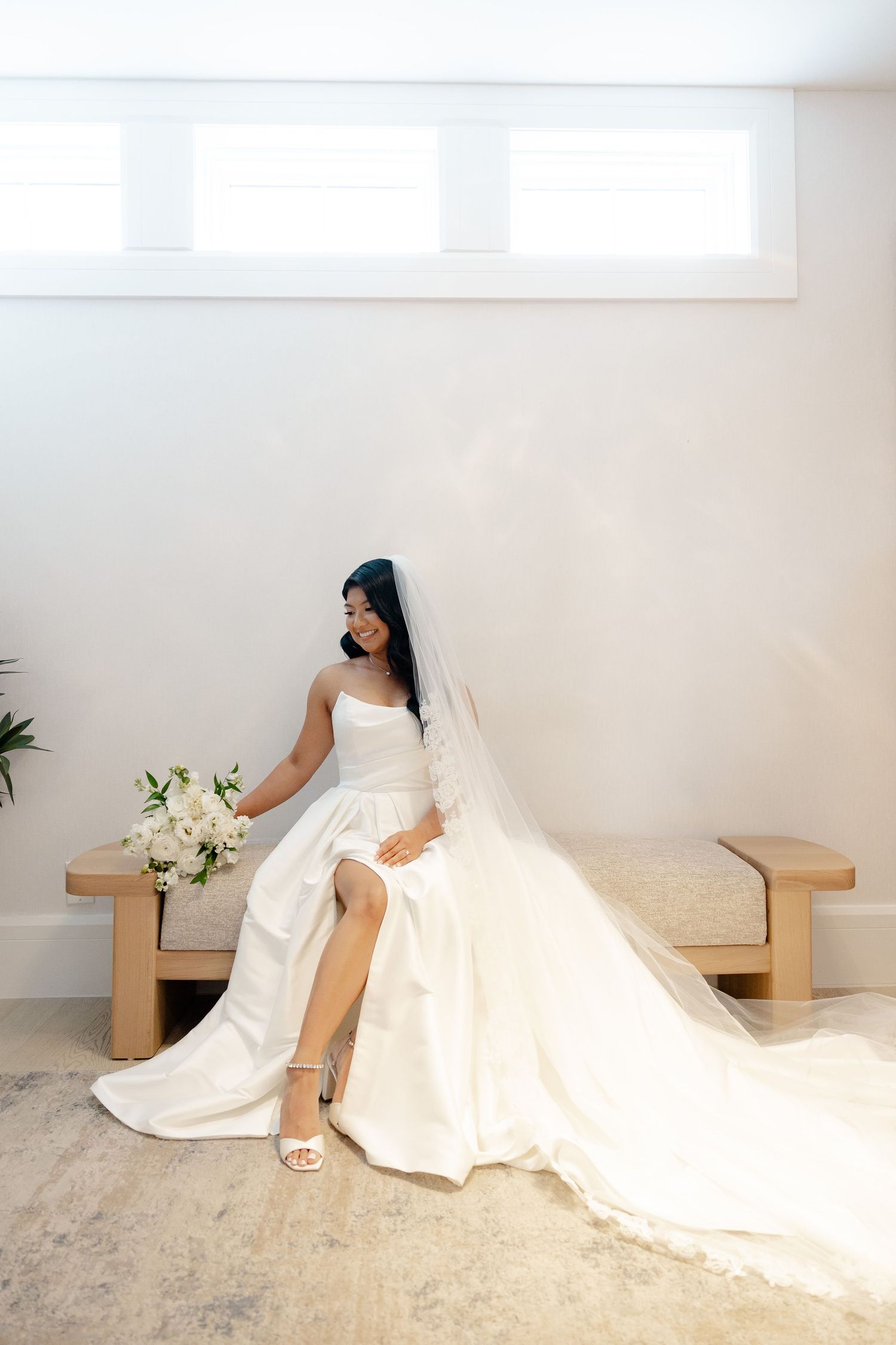 Bride in an elegant white gown sits on a bench showing off her dress and veil.