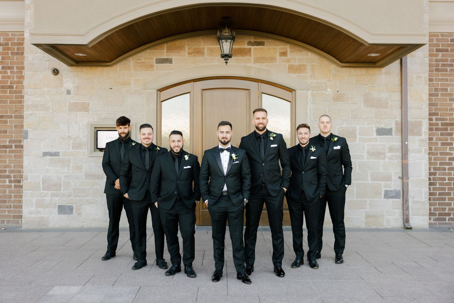 Wedding party members pose together in black suits against a beige stone building backdrop.