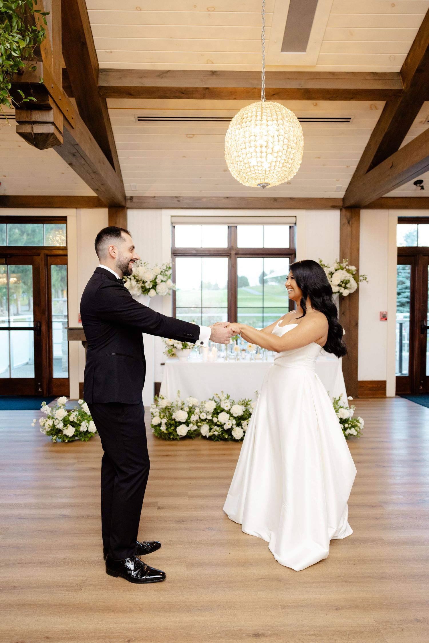 Couple shares a dance in sunlit reception venue with rustic wooden beams and crystal lighting.