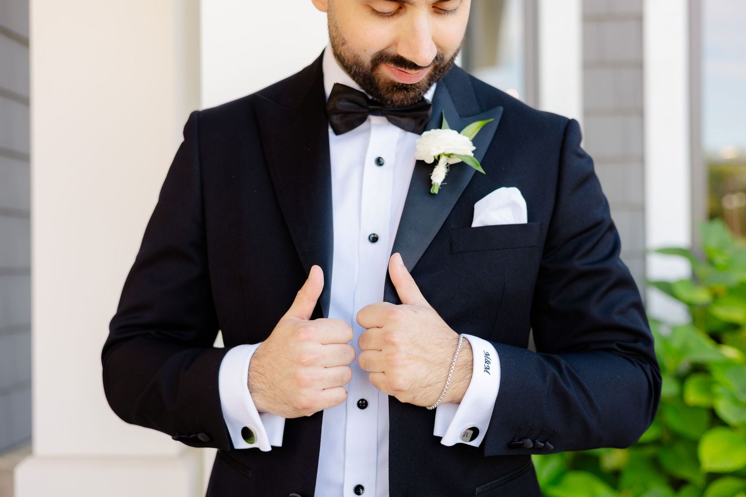A close-up detail shot of a groom adjusting his black tuxedo with white boutonniere and bowtie.