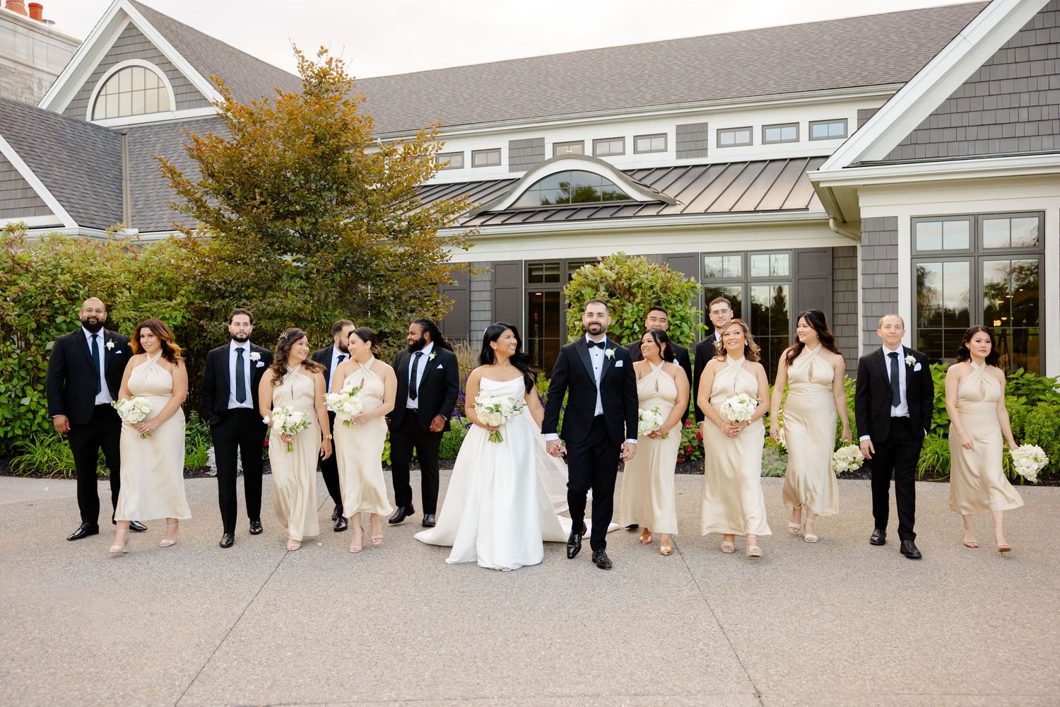 Wedding party in black and cream attire poses casually outside coastal-style venue.