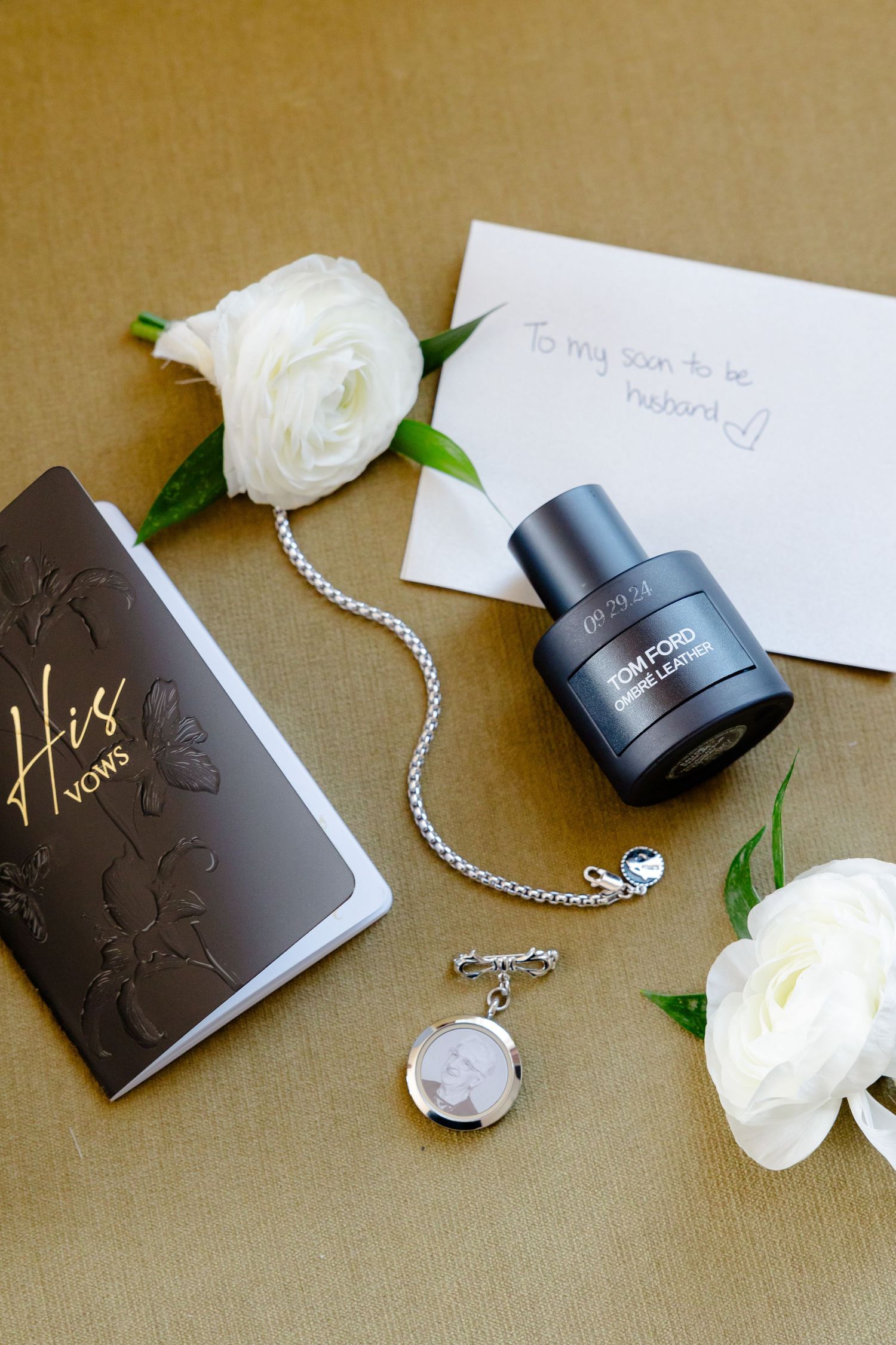 Wedding details flat lay with cologne, jewelry, white roses and note on brown surface.