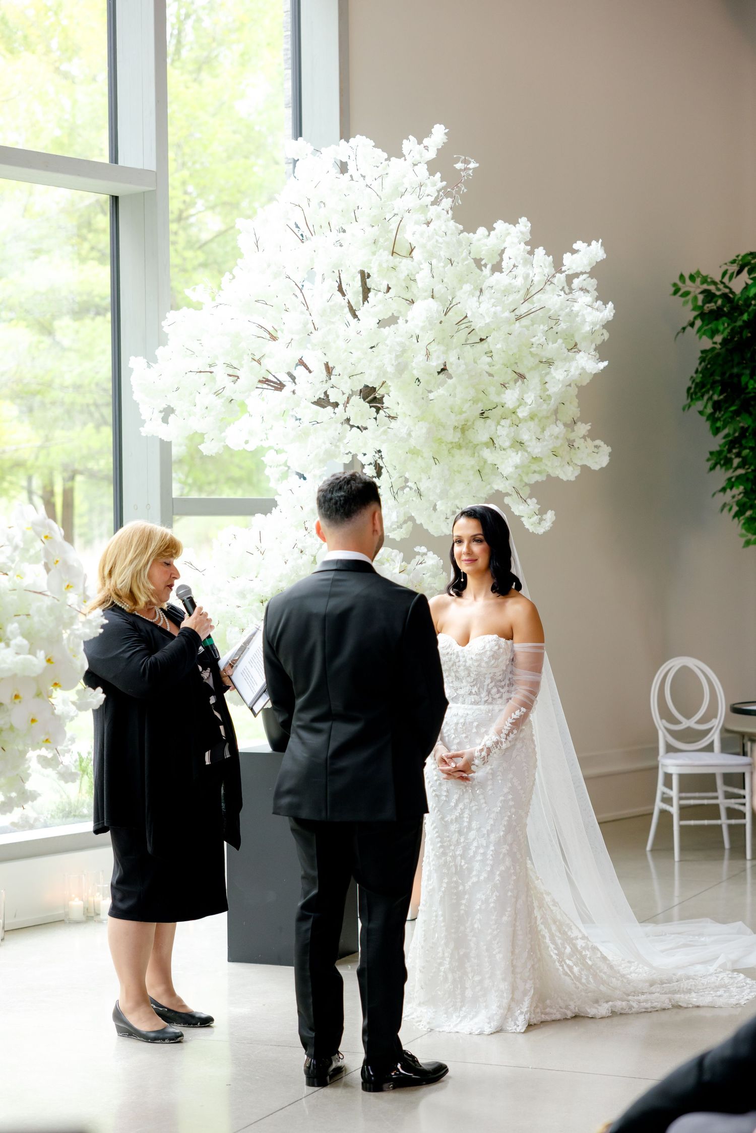 An elegant wedding ceremony with white floral decor and natural lighting from floor-to-ceiling windows.
