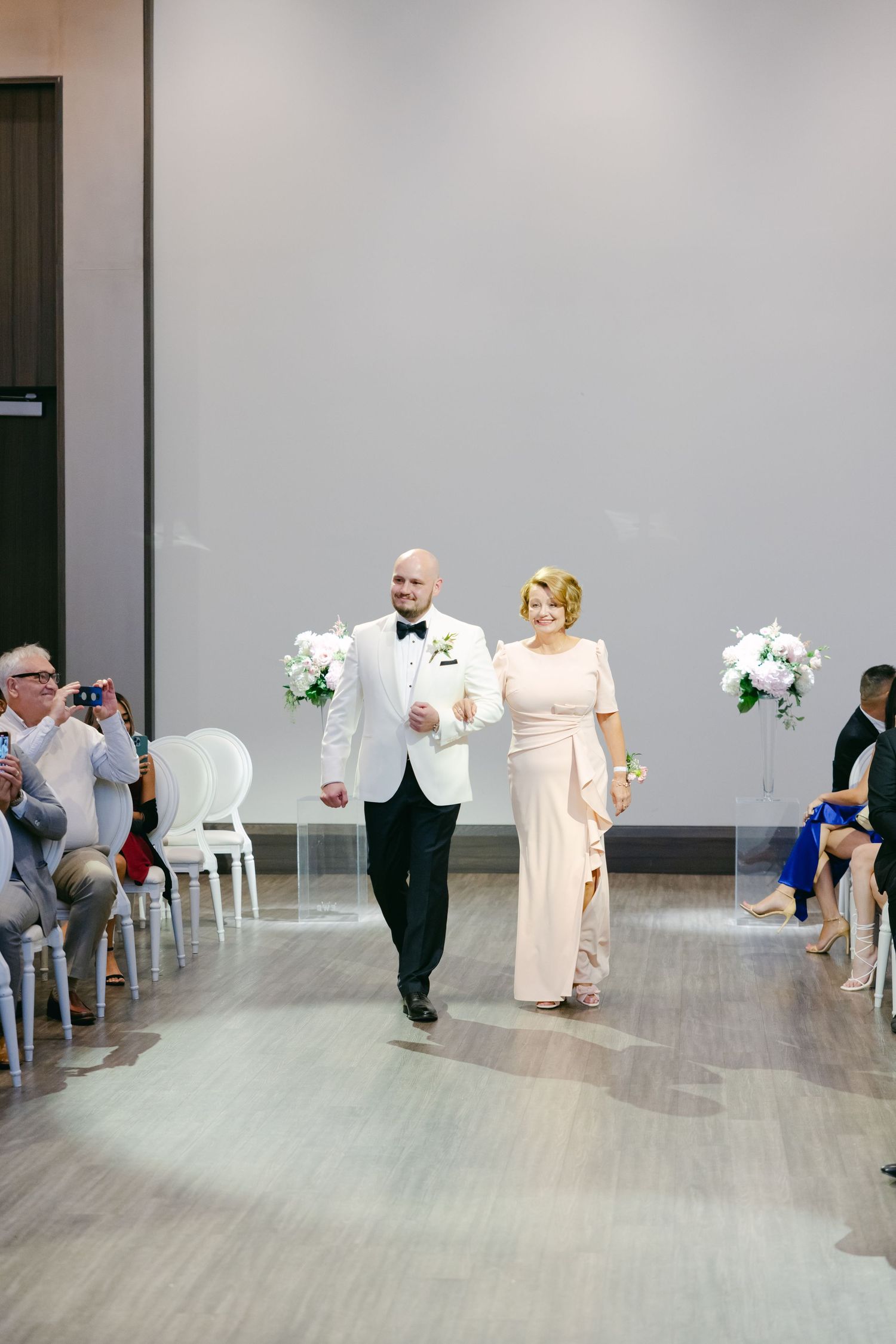A couple walking down the aisle at a wedding ceremony with white floral arrangements in an elegant indoor venue.