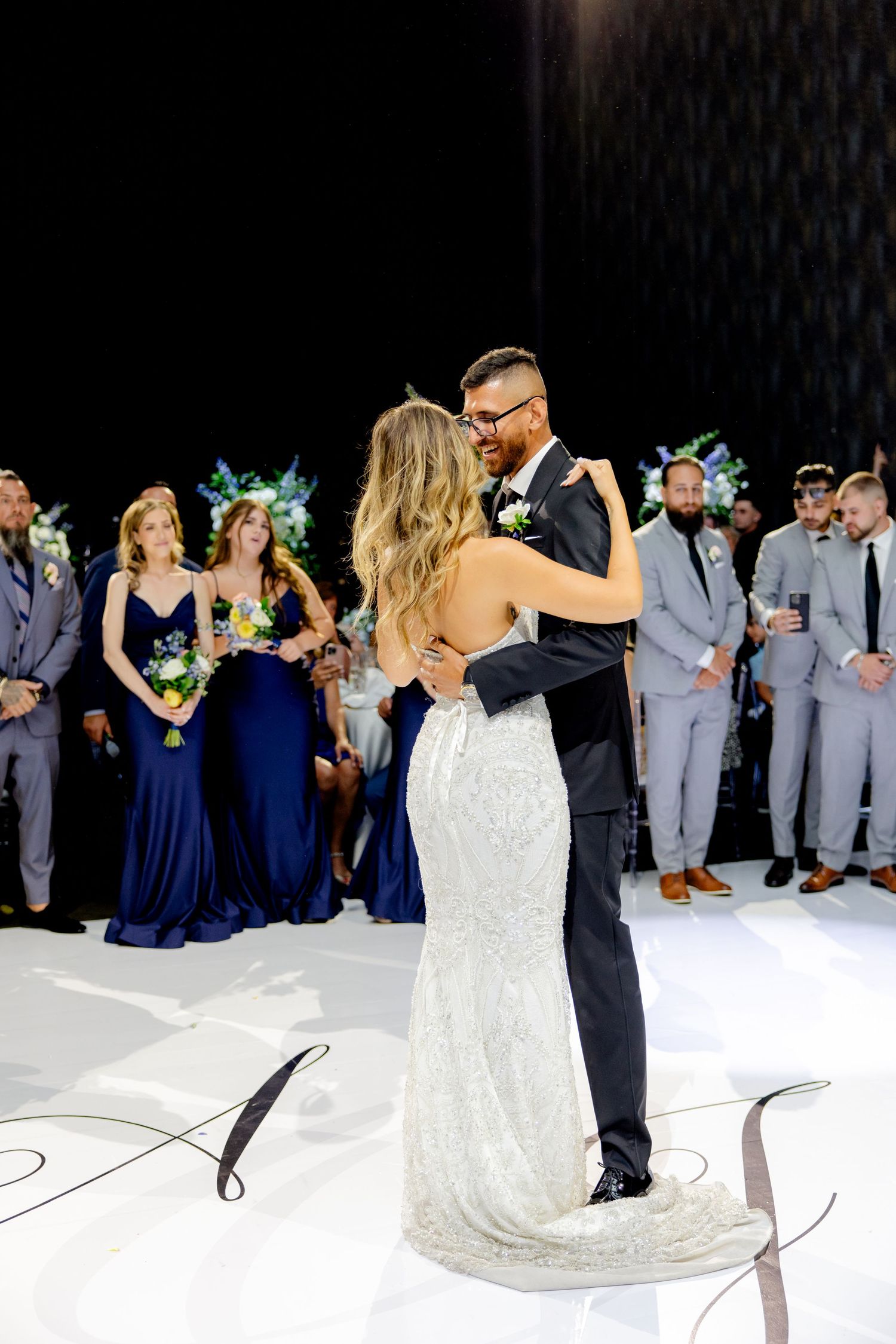 Couple shares their first dance while wedding party watches from the sidelines.