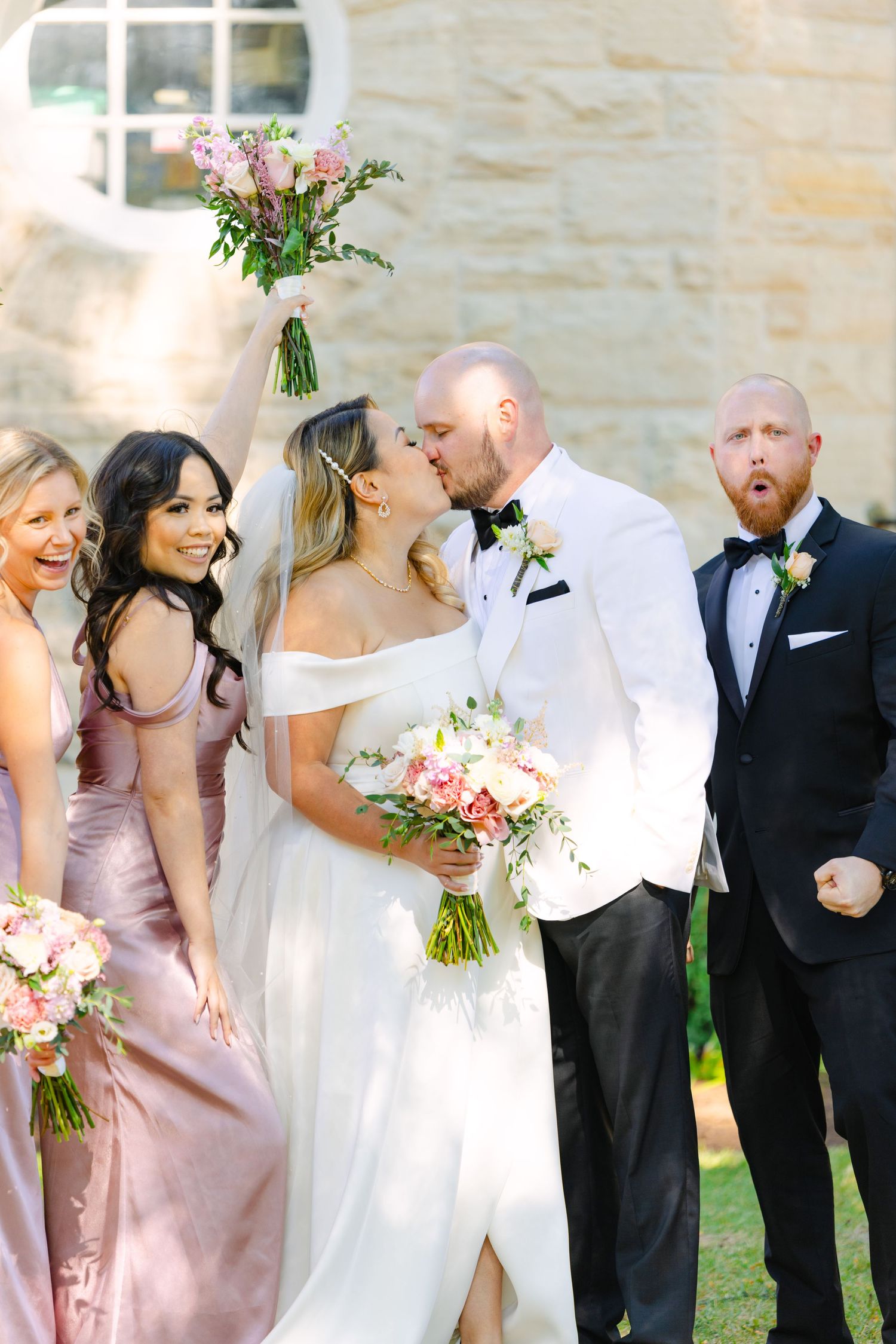 Wedding party celebrating with the newlyweds sharing a kiss in front of a stone church.