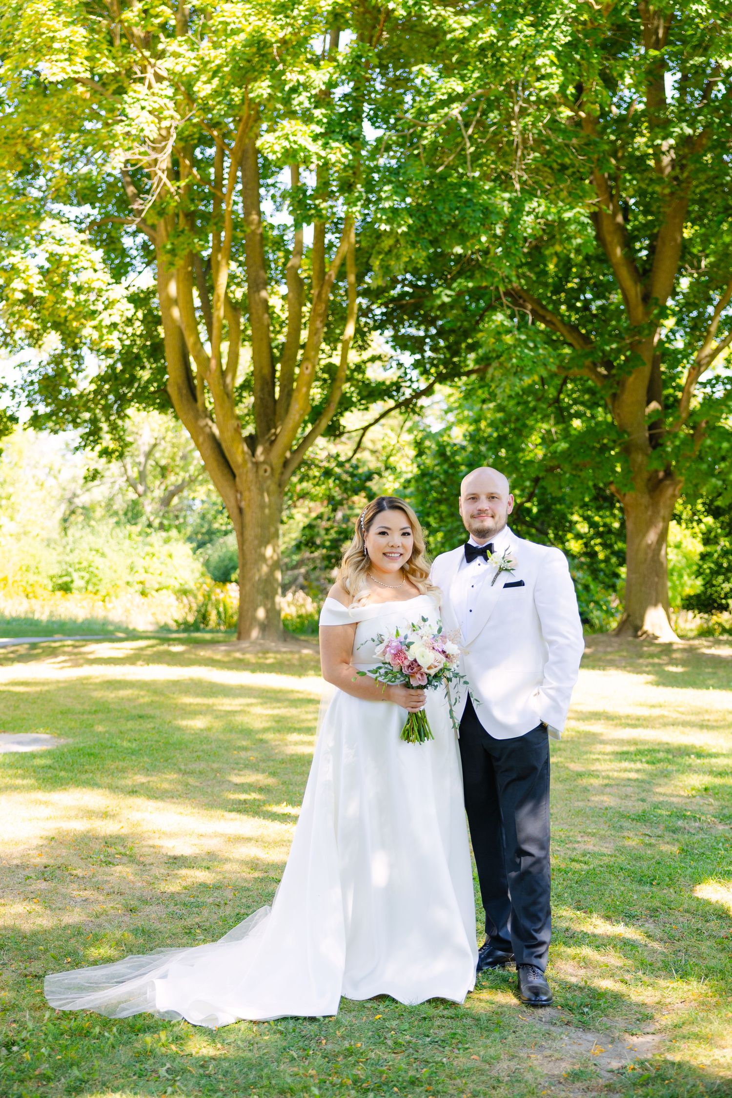 Wedding couple poses outdoors under large trees in a garden setting with natural sunlight filtering through.